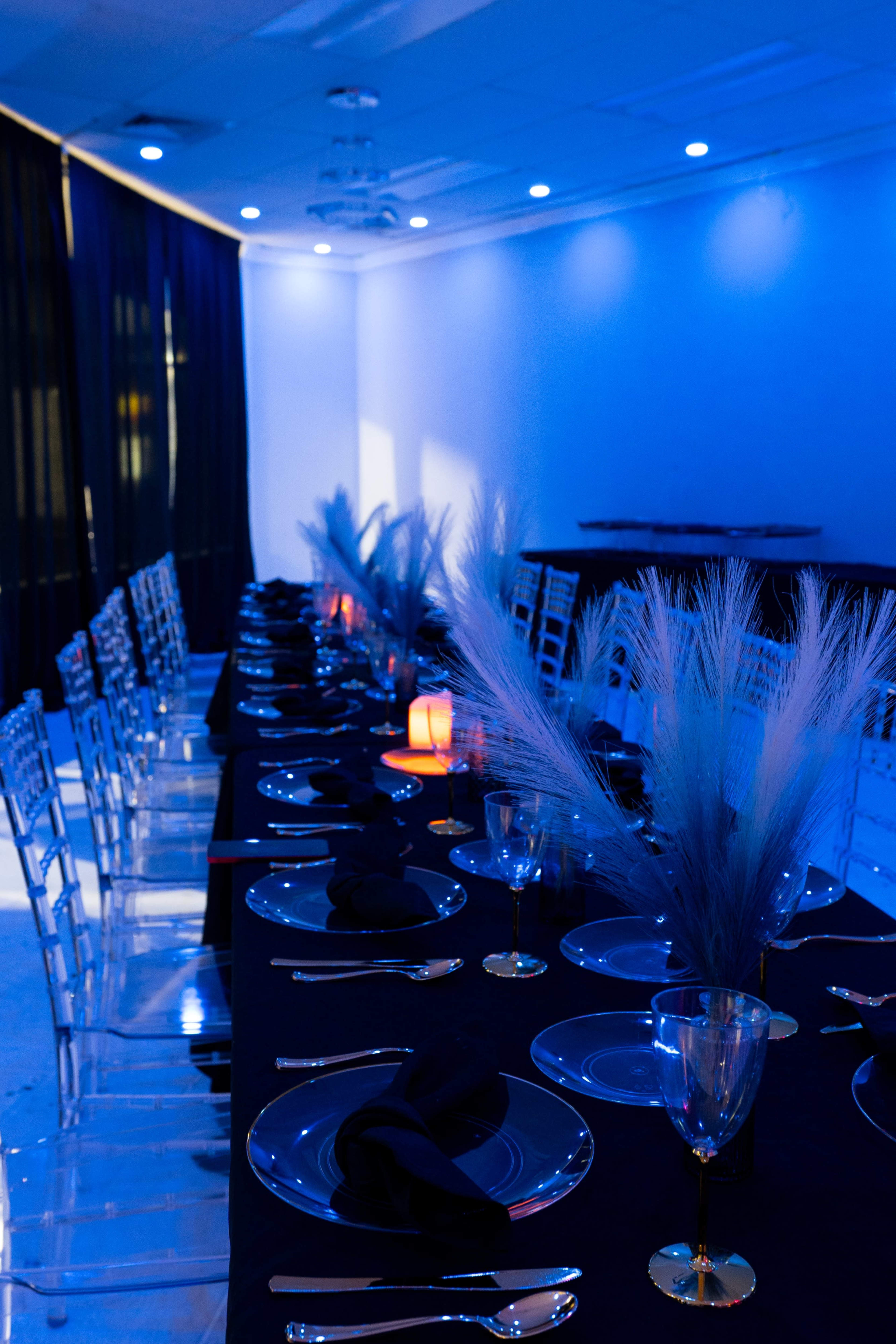 A long dining table is set with black tablecloths, clear plates, elegant glassware, and decorative pampas grass, illuminated by blue lighting.