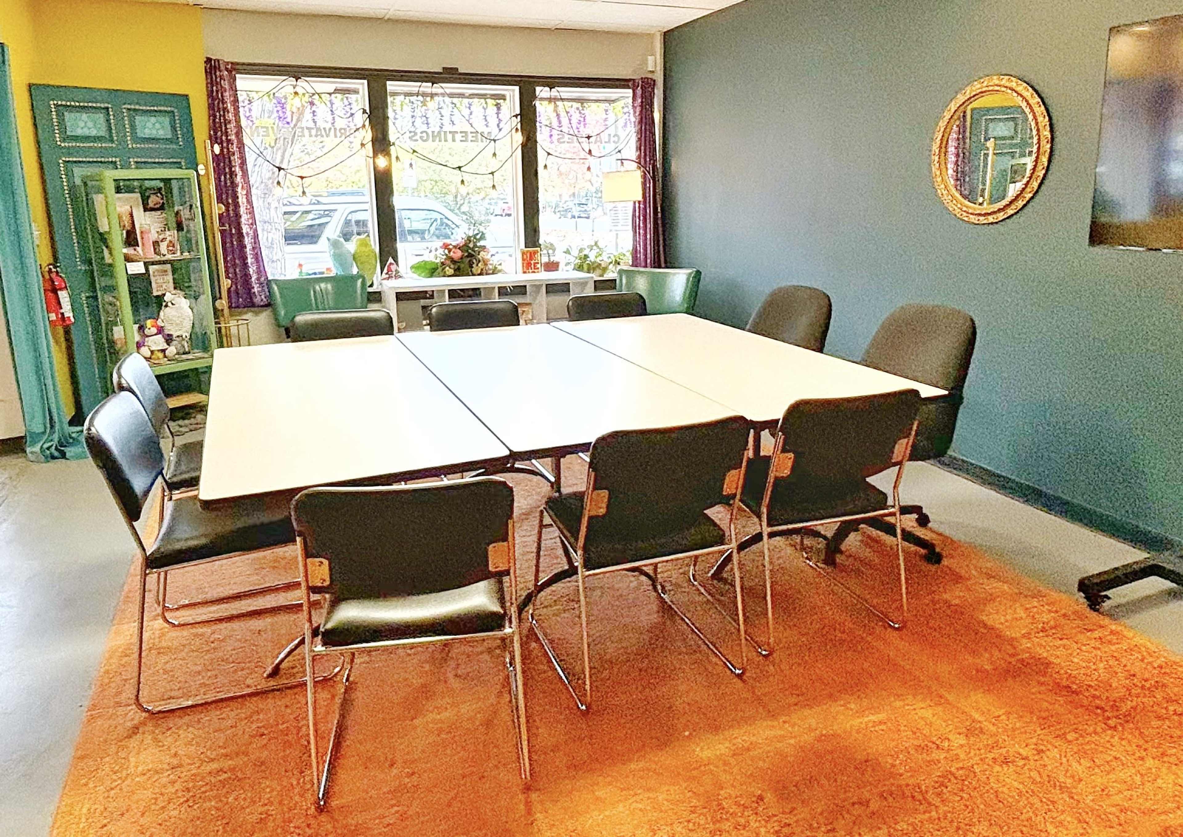 A meeting room features a long table surrounded by black chairs, with colorful decorations and a large mirror on the wall.