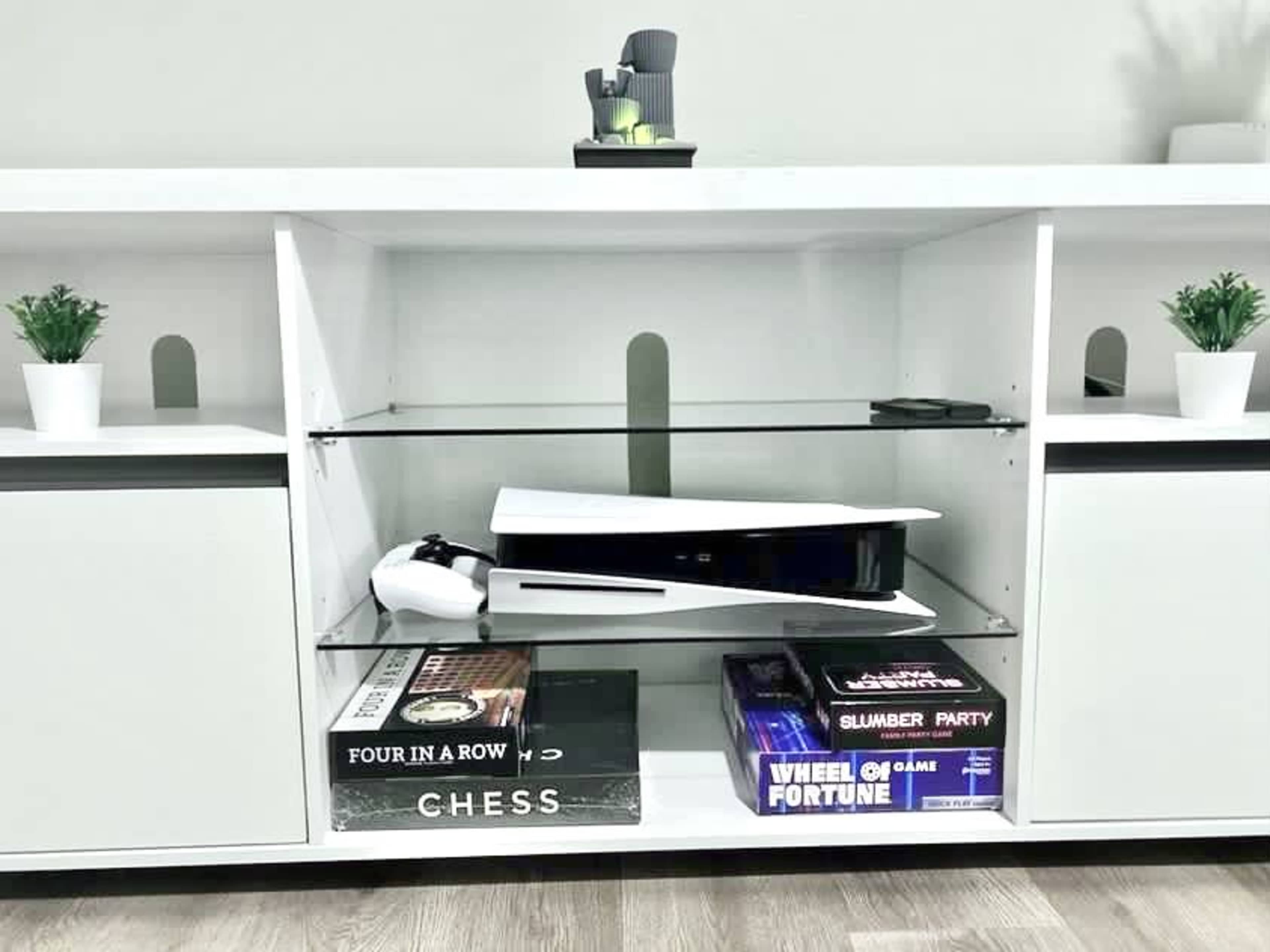 A modern console table displays a PlayStation 5, board games, and small potted plants on its shelves.