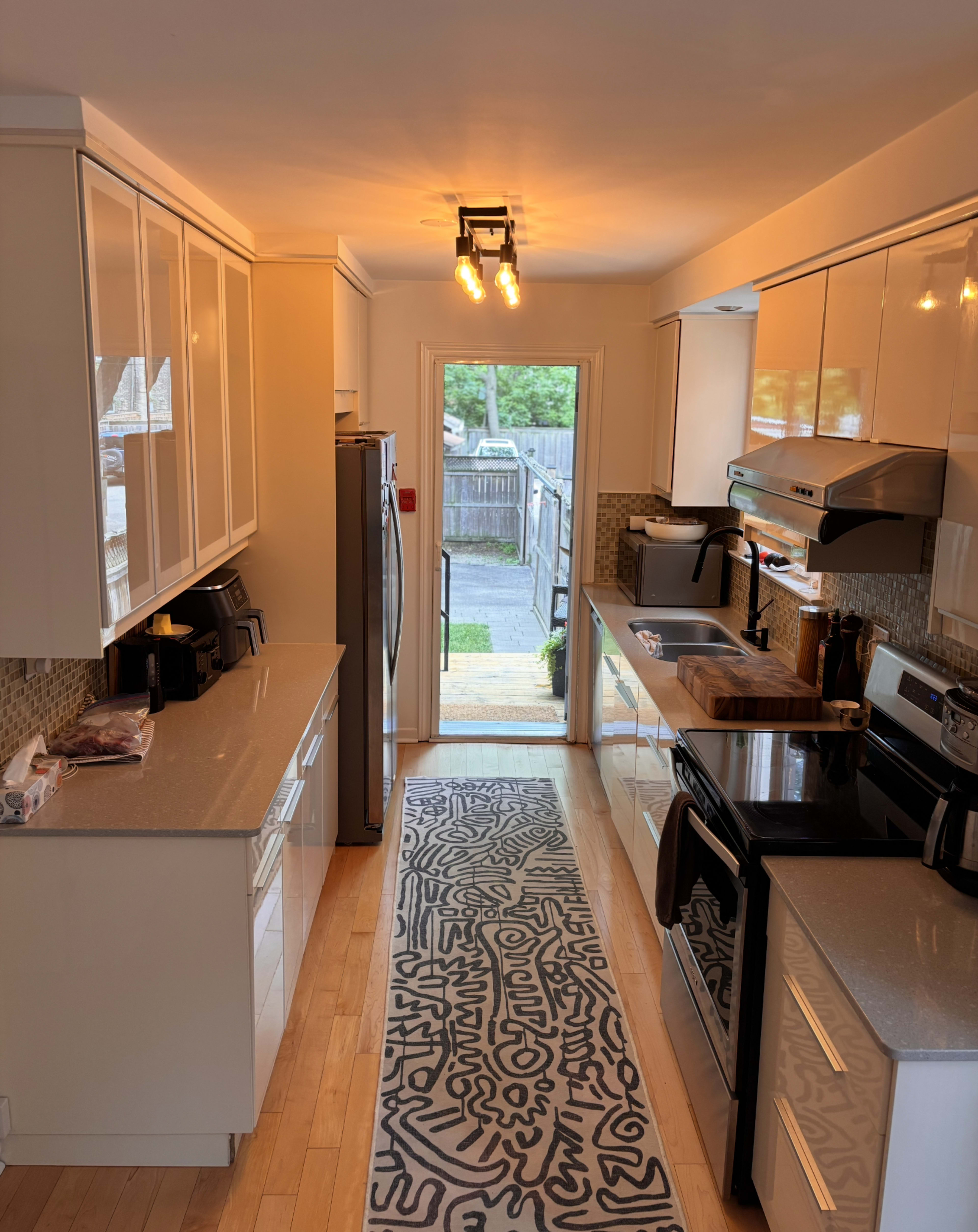 The image shows a modern kitchen with white cabinetry, a gray countertop, stainless steel appliances, and a door leading to an outdoor area.