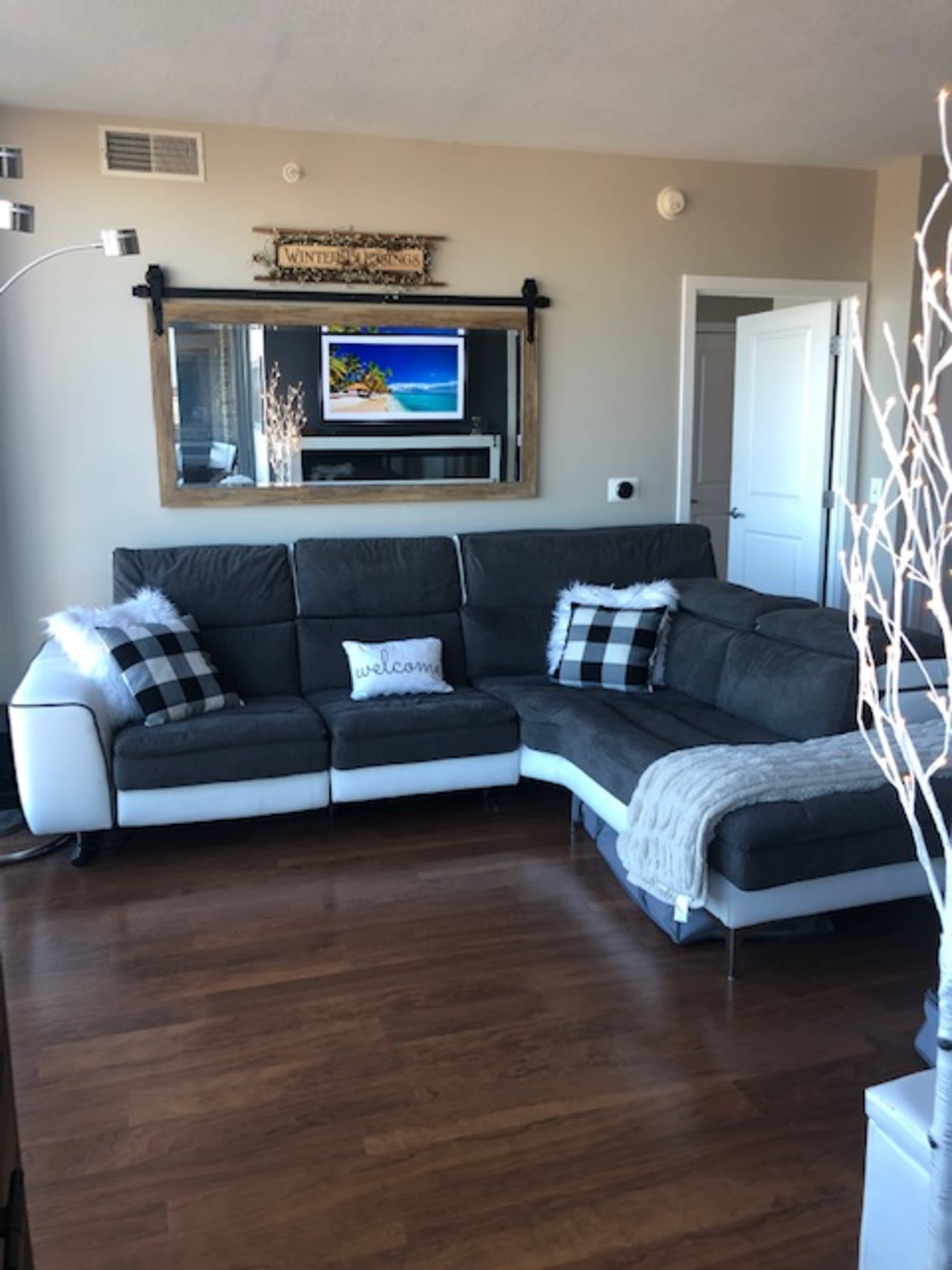 A living room with a gray sectional sofa, a large mirror above a TV, and a decorative welcome pillow.
