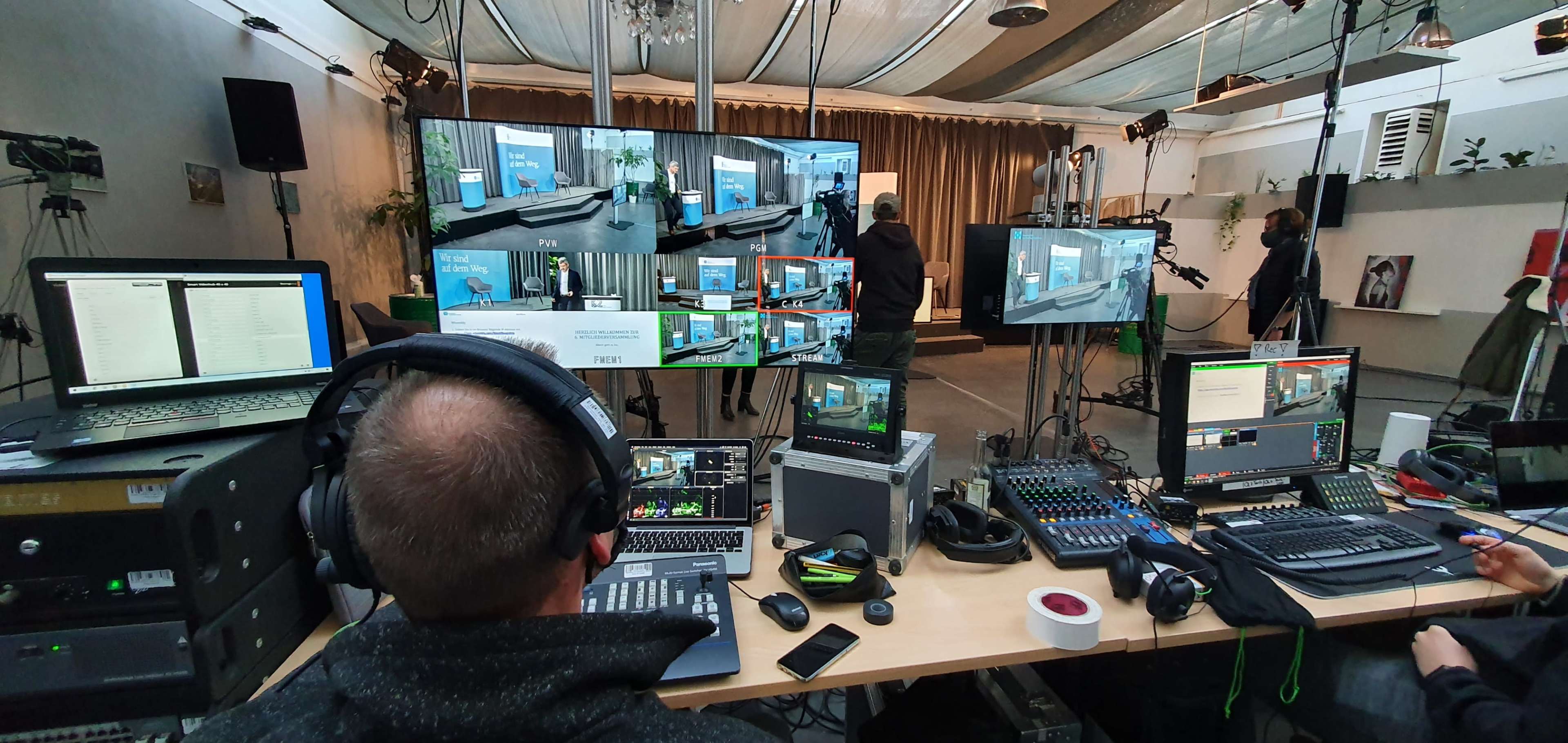 A man operates audio-visual equipment at a control station with multiple screens in a studio setting.