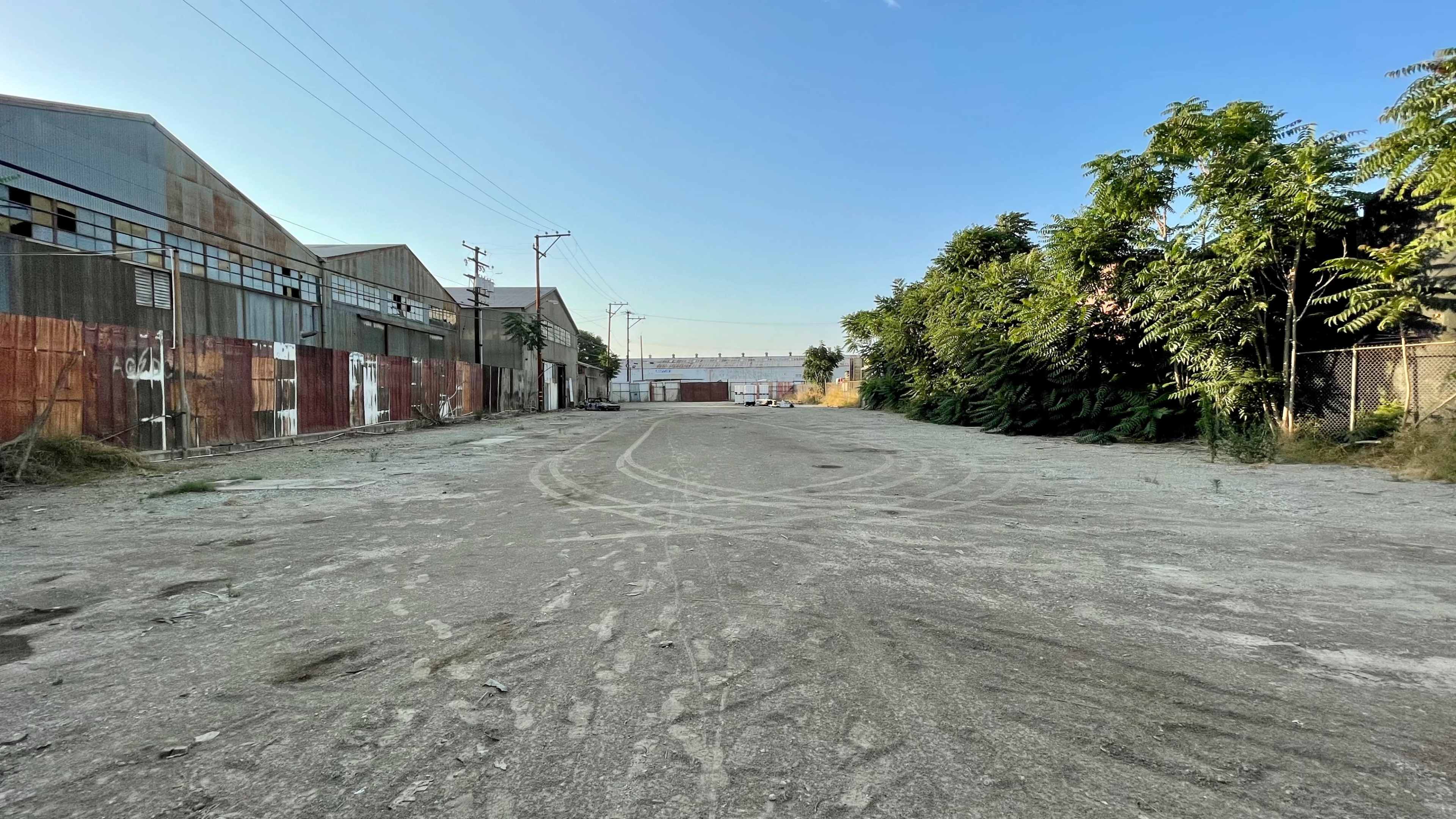An empty, gravel road runs through an industrial area with rusted metal buildings and overgrown foliage on either side.