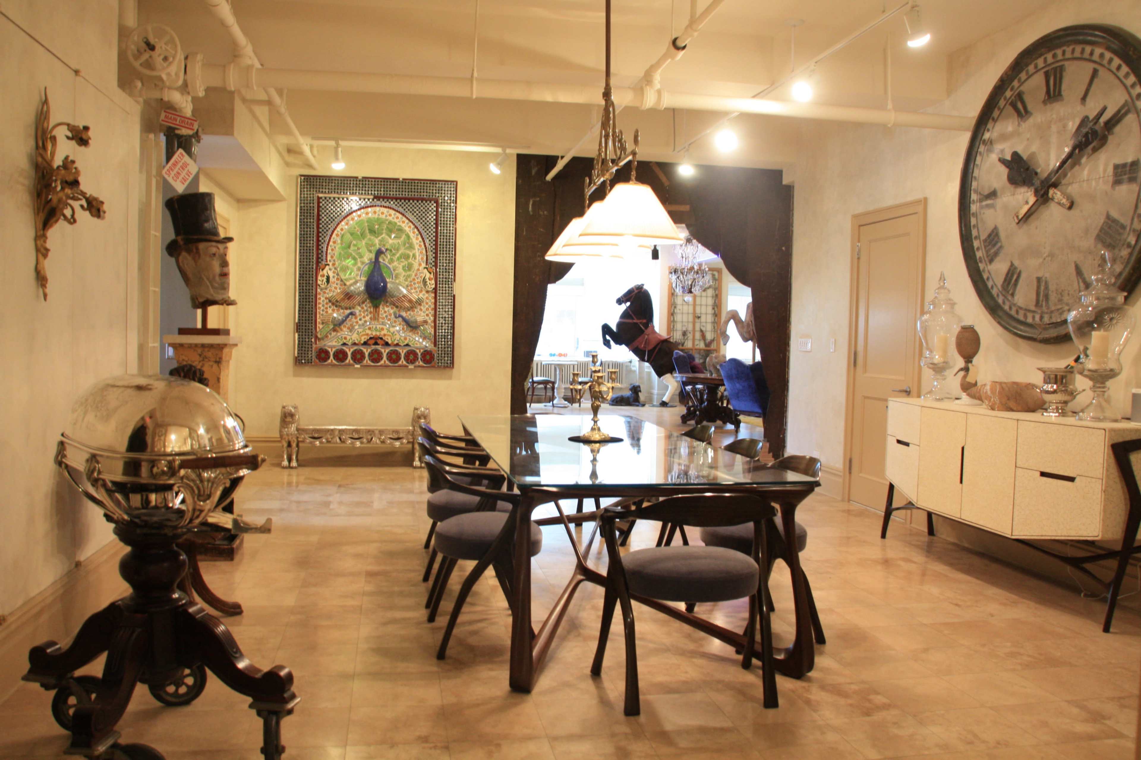 The image shows a modern dining area with a glass table surrounded by chairs, sculptures, and a large decorative clock on the wall.