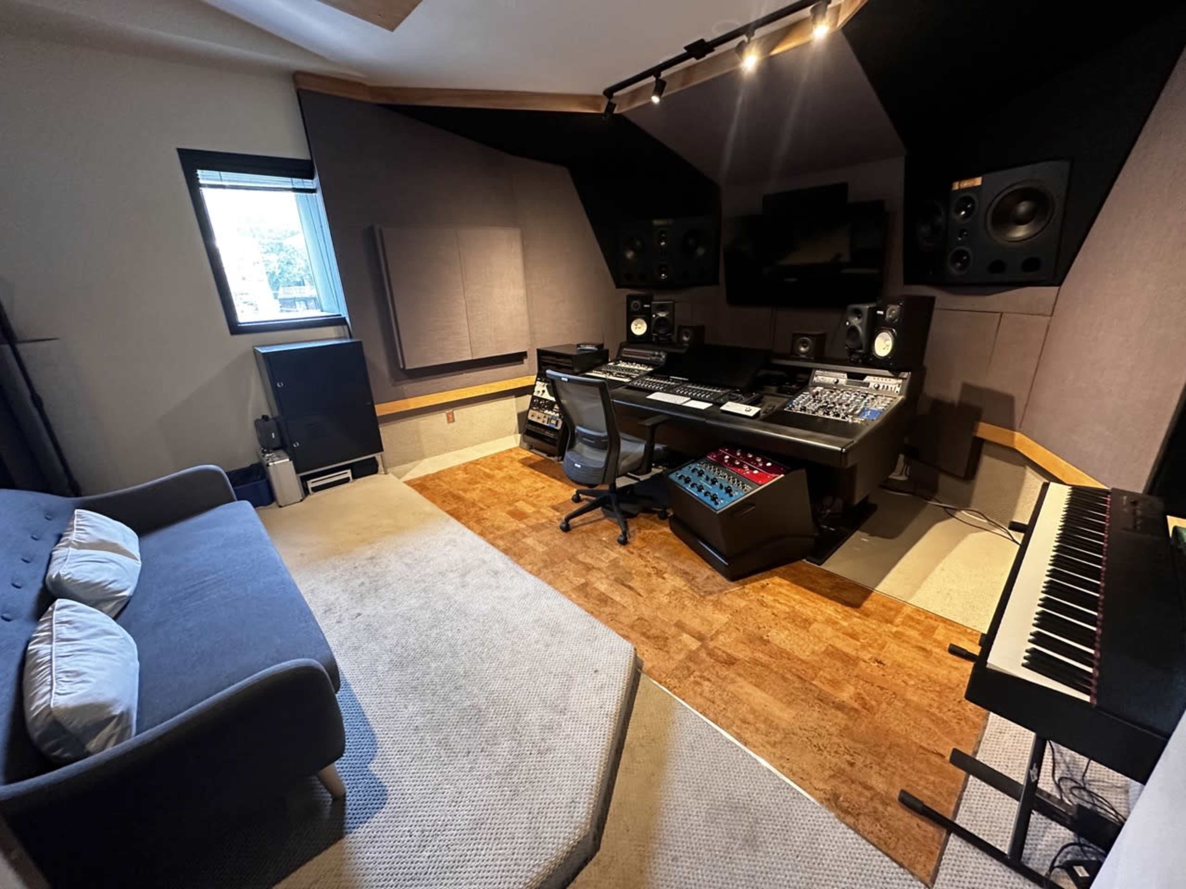 The image shows a music studio featuring a mixing desk with various audio equipment, large speakers, a keyboard, and a couch in the corner.