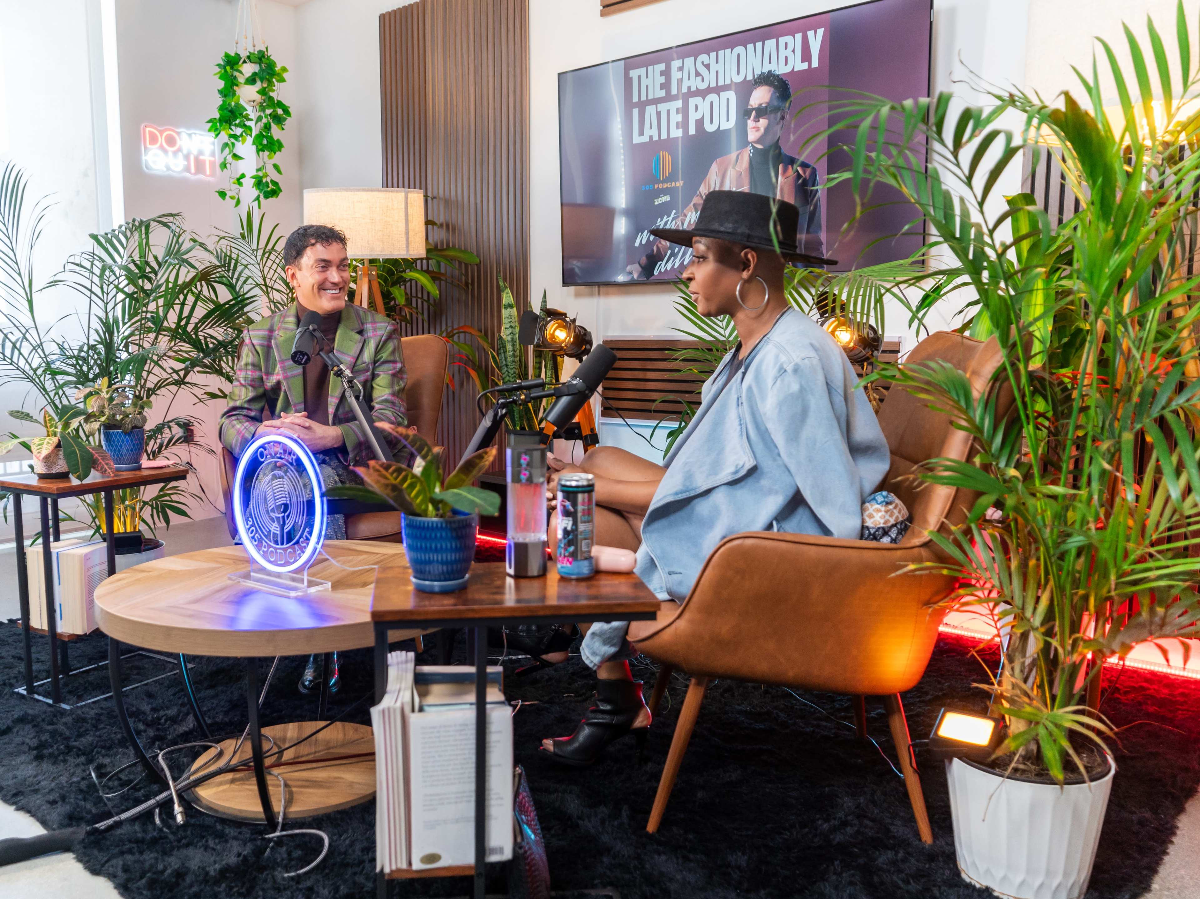 A man and a woman sit in a brightly lit studio with plants and a large screen displaying the title "The Fashionably Late Pod."