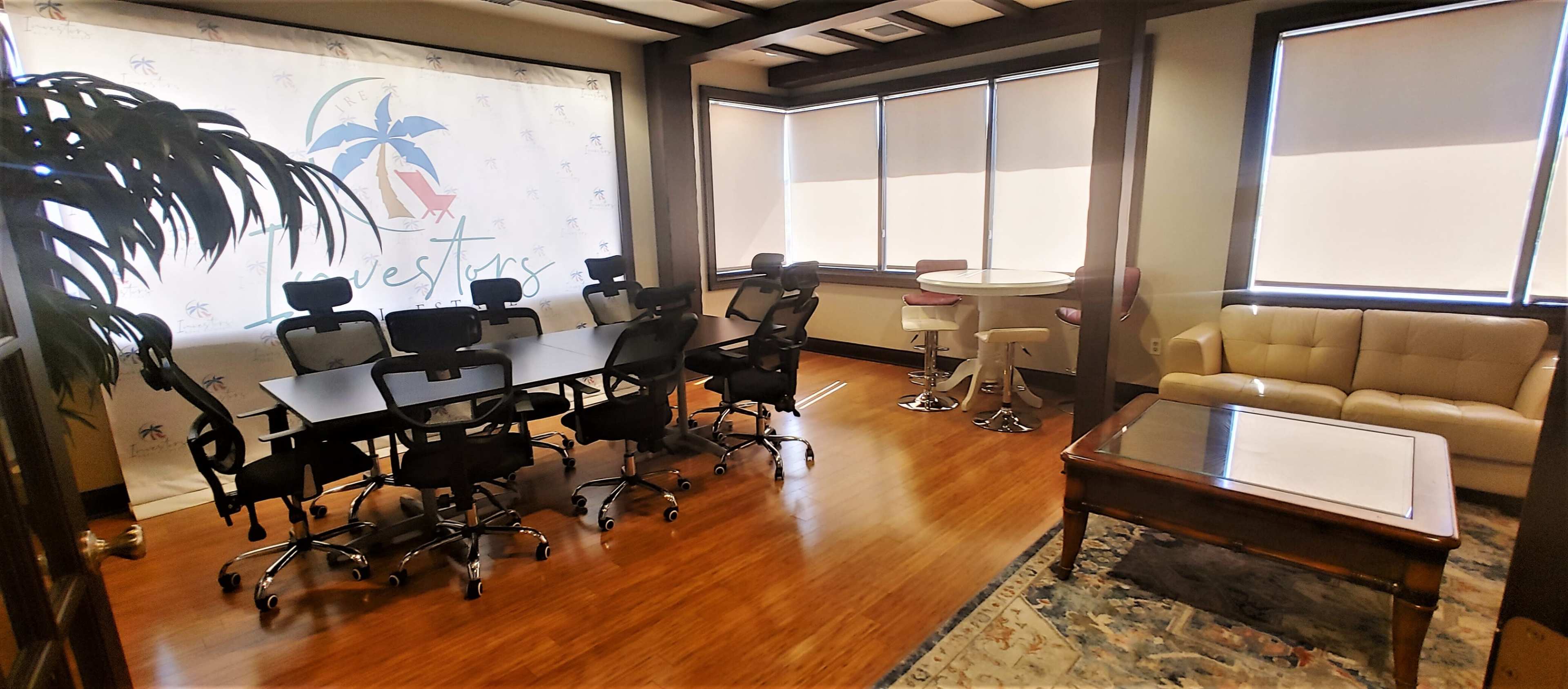 A conference room features a large table with black chairs, a round bar table with stools, a beige lounge couch, and a backdrop displaying a logo with palm trees.