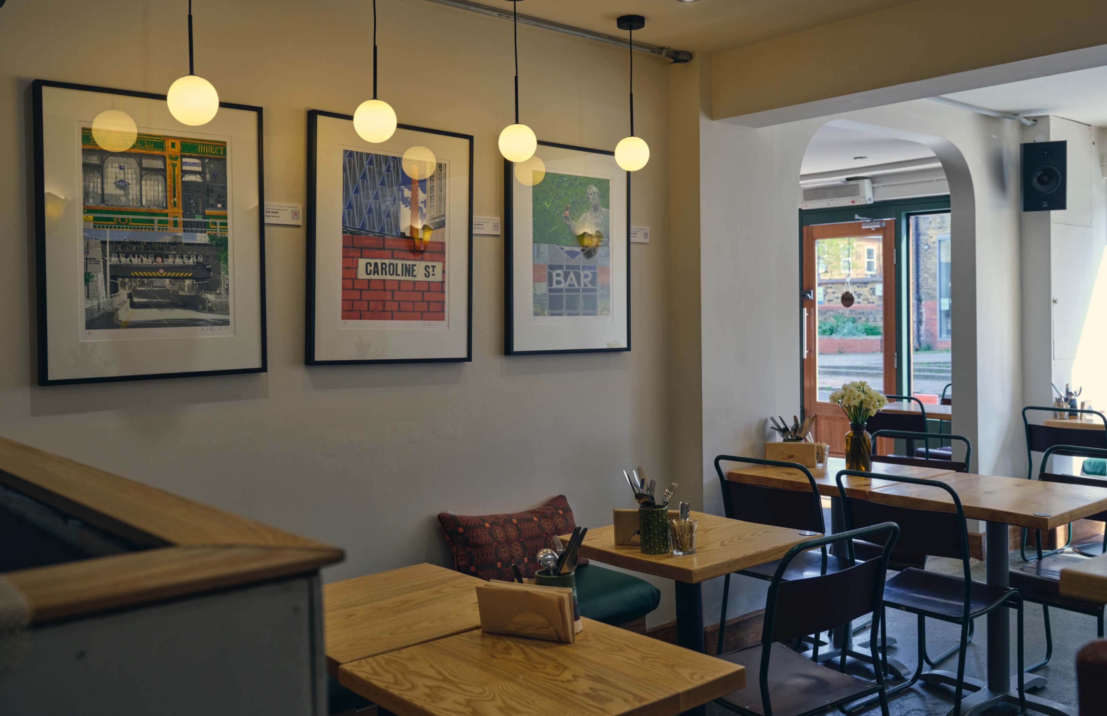 The image shows a cozy café interior with wooden tables and framed artwork on the walls, illuminated by pendant lights.