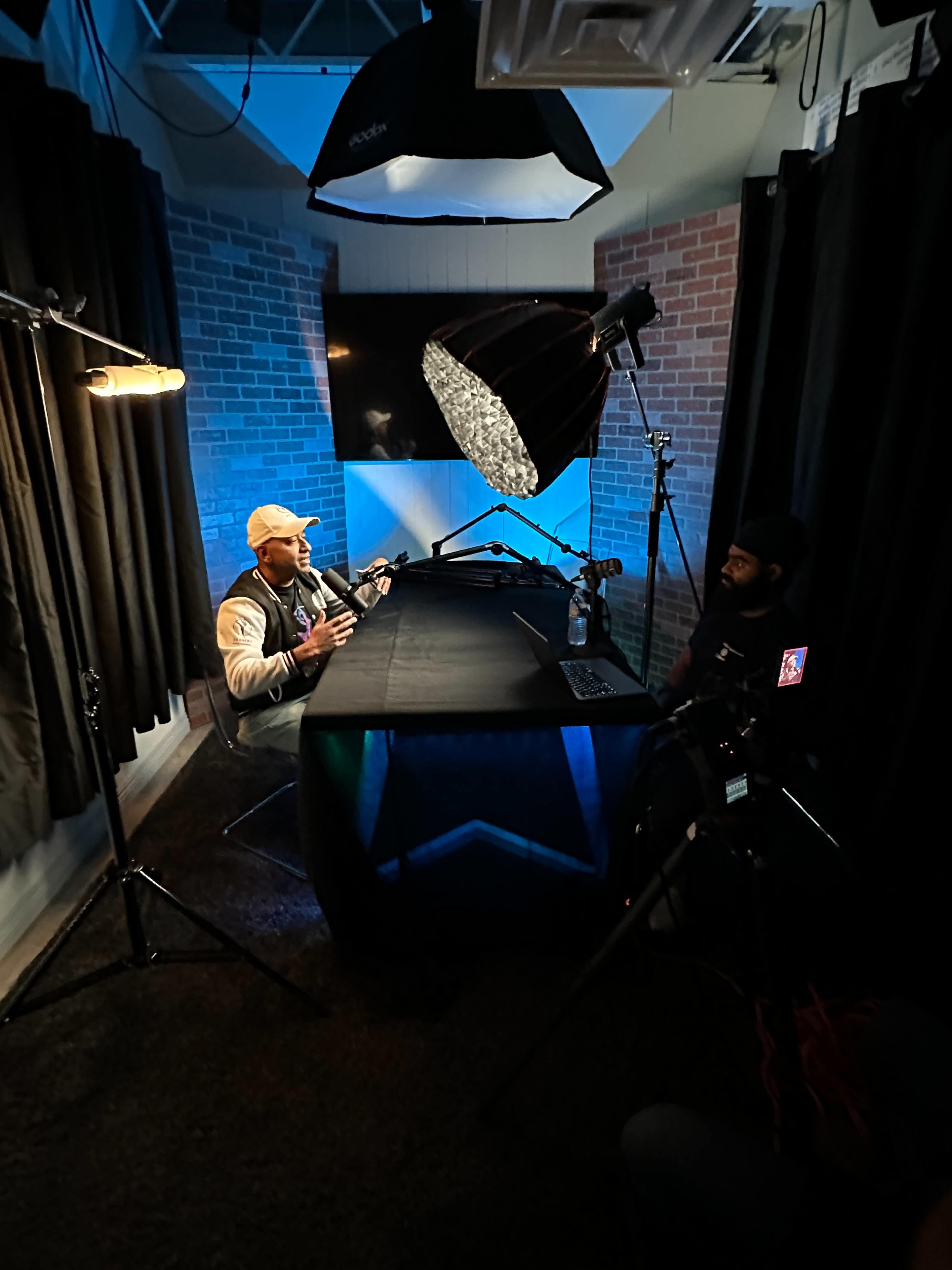 A person is sitting at a table under a large light fixture in a recording studio with dark curtains and a brick wall backdrop.