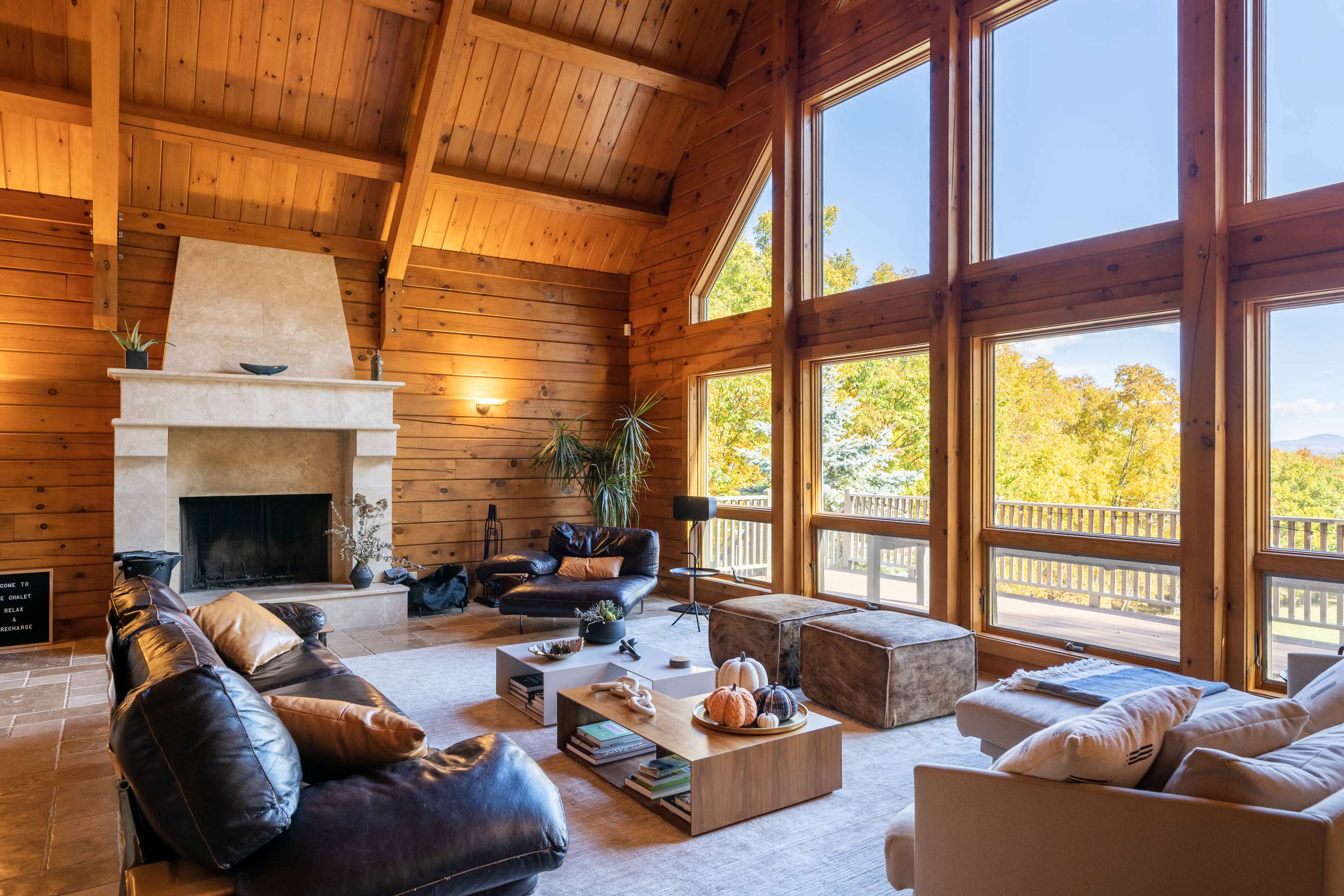 The interior of a wooden cabin features a spacious living area with large windows overlooking trees and a fireplace at one end.