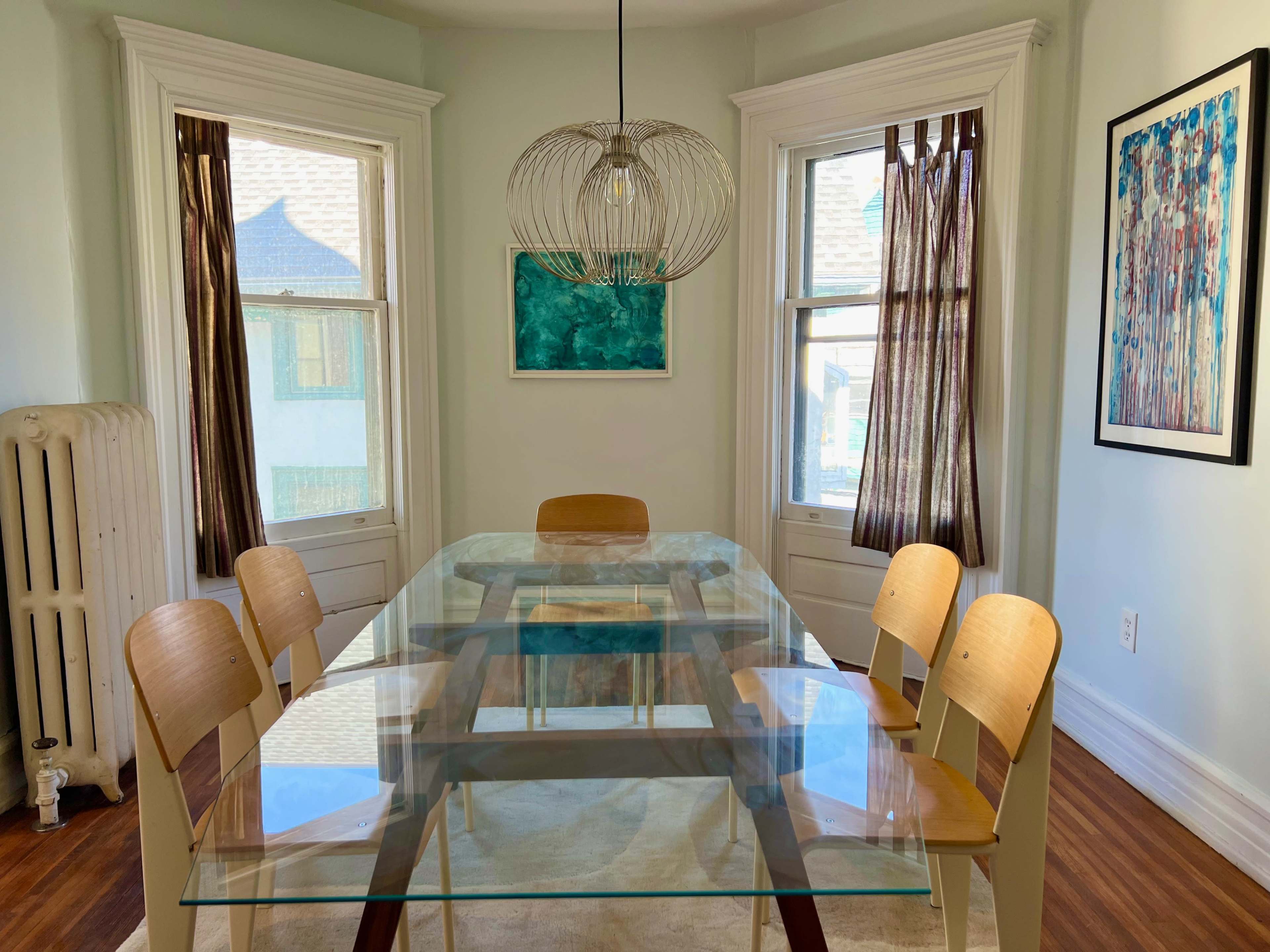 A modern dining room features a glass table surrounded by wooden chairs, with large windows and a hanging light fixture above.