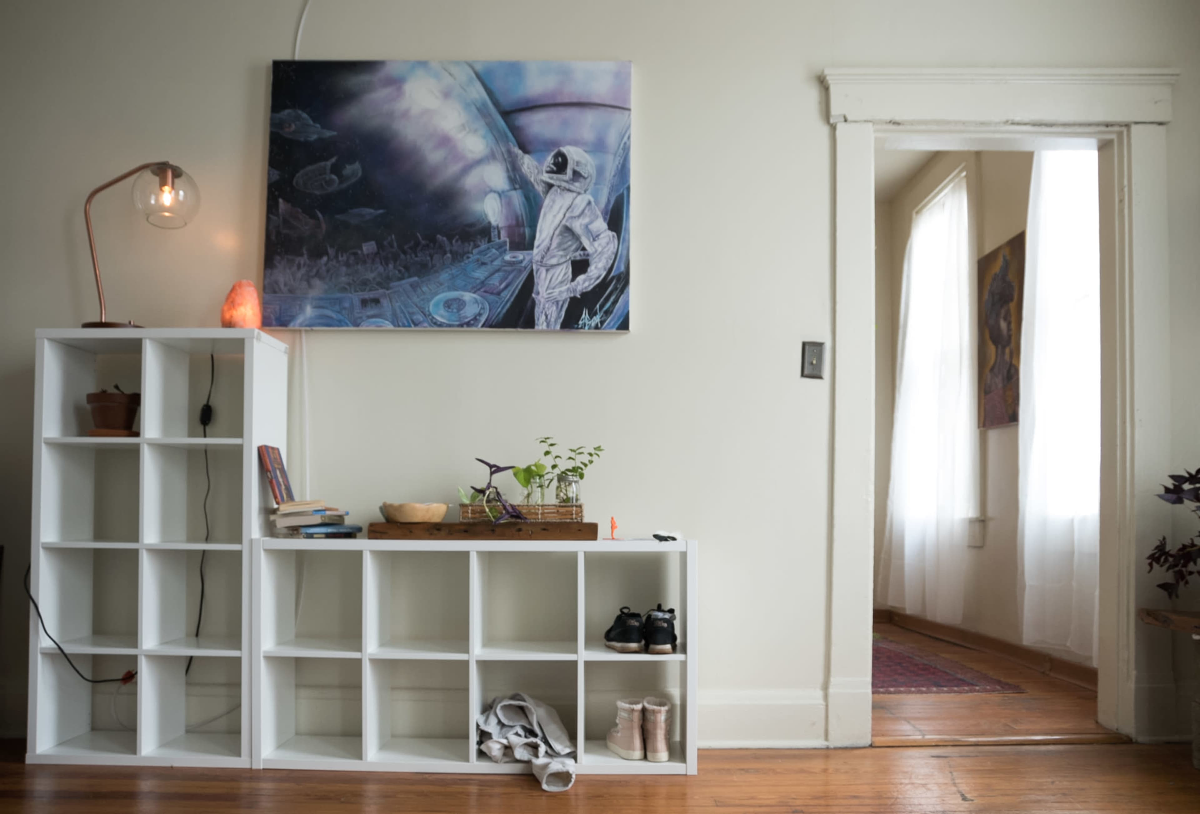 The image shows a bright room with a white bookshelf displaying plants and books, and a painting of an astronaut hanging on the wall, with a doorway leading to another room.