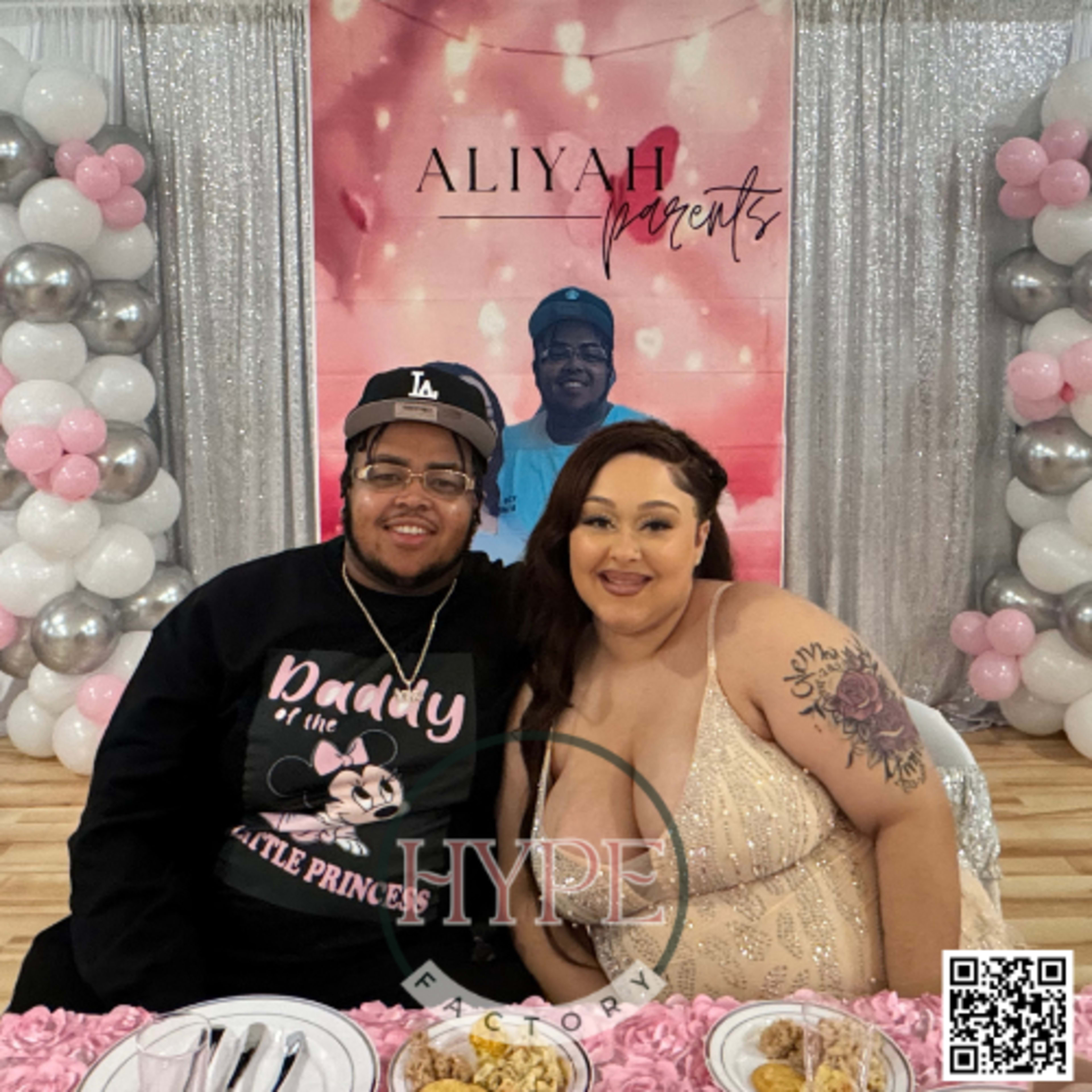 A couple sits at a decorated table with a large backdrop and balloons in a celebratory setting.