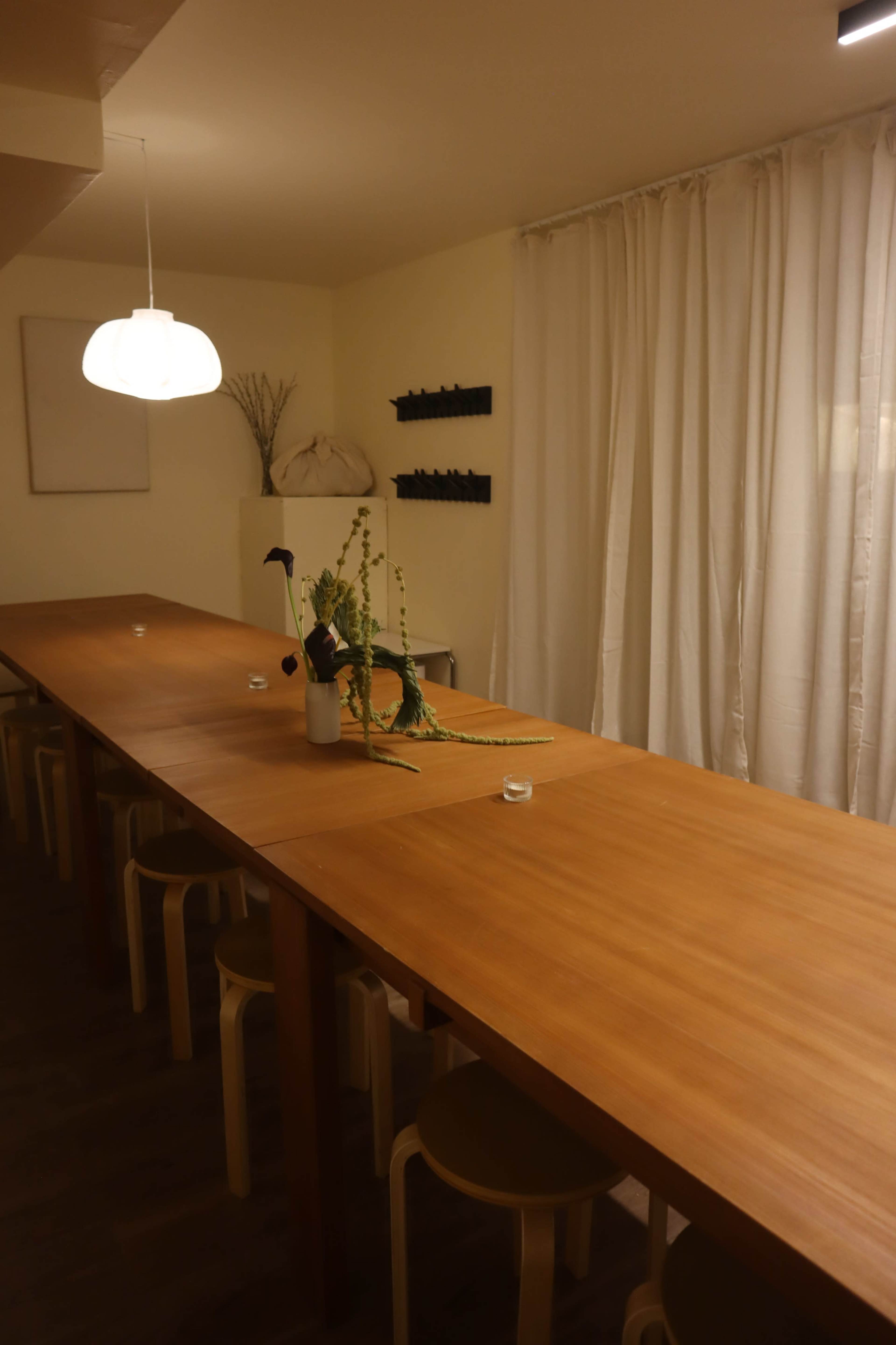 A long wooden table with several chairs is set in a softly lit room featuring white curtains and simple decor.