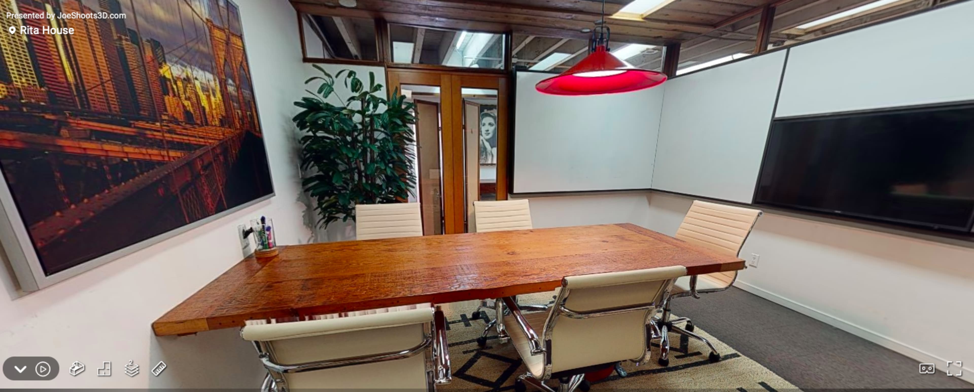 A modern conference room features a large wooden table surrounded by white leather chairs, with a red pendant light and wall-mounted displays.
