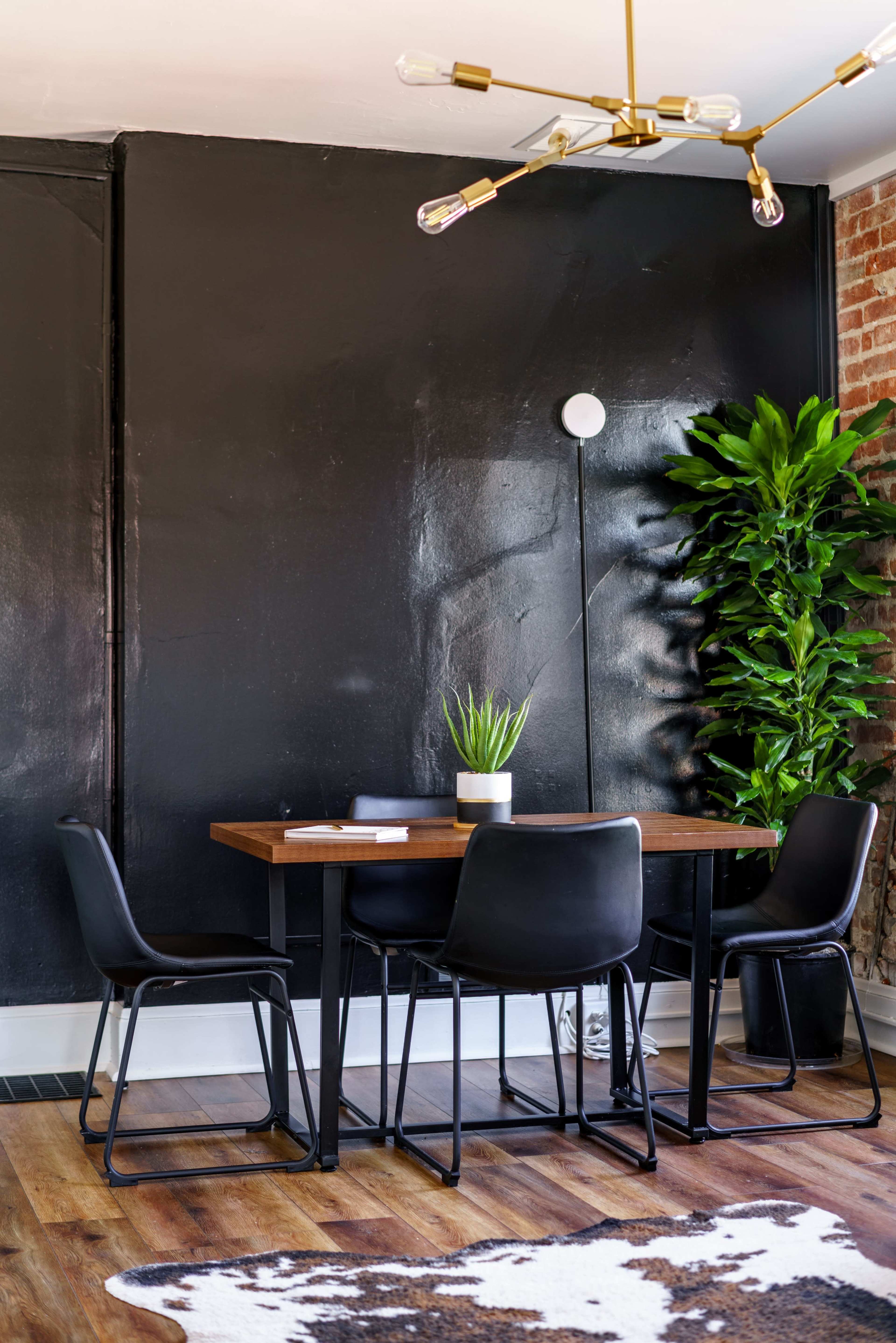 The image shows a modern dining area with a wooden table surrounded by four black chairs, a large potted plant in the corner, and a black wall behind.