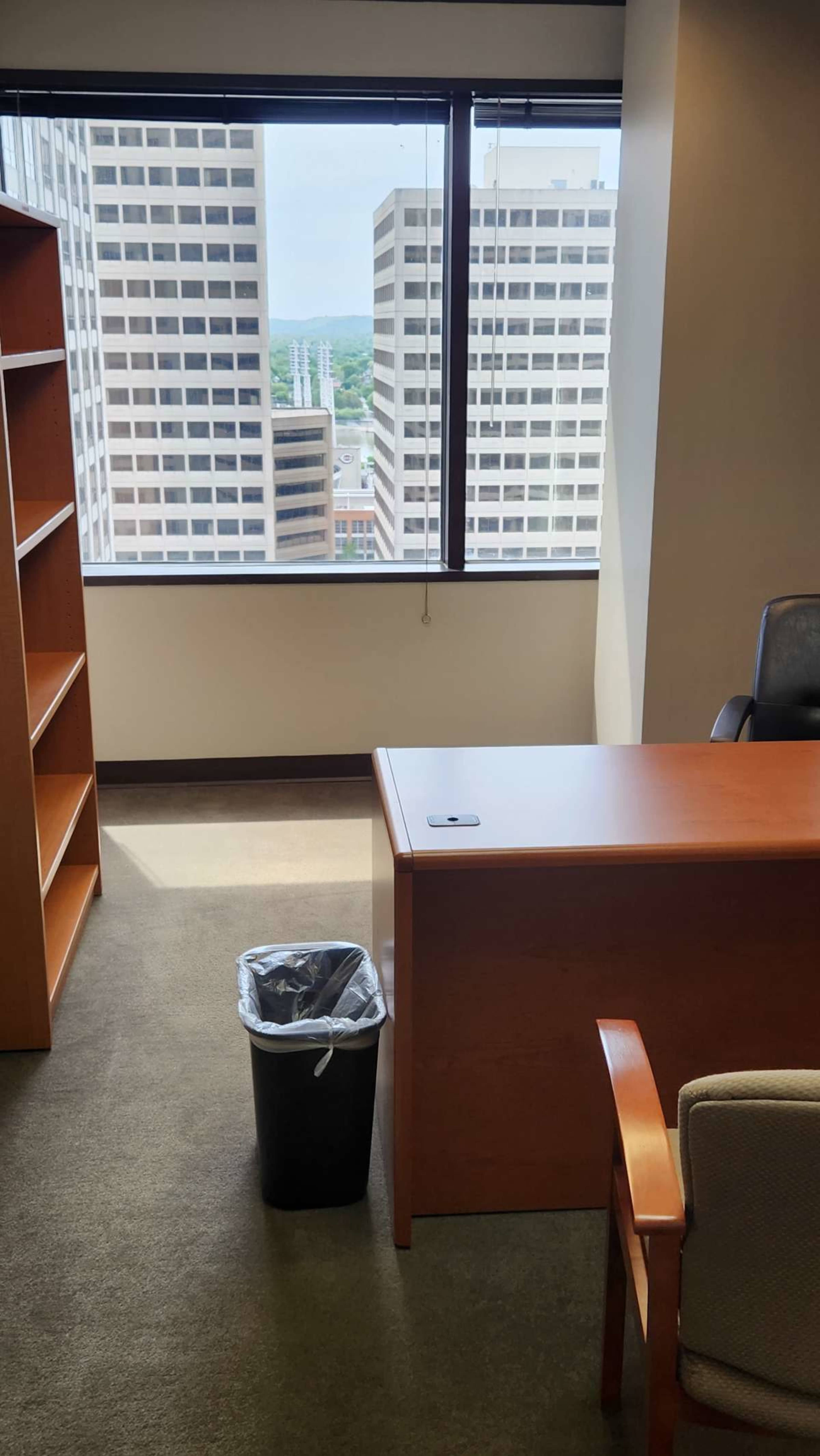 A modern office space with a wooden desk, a chair, a wastebasket, and large windows overlooking high-rise buildings.