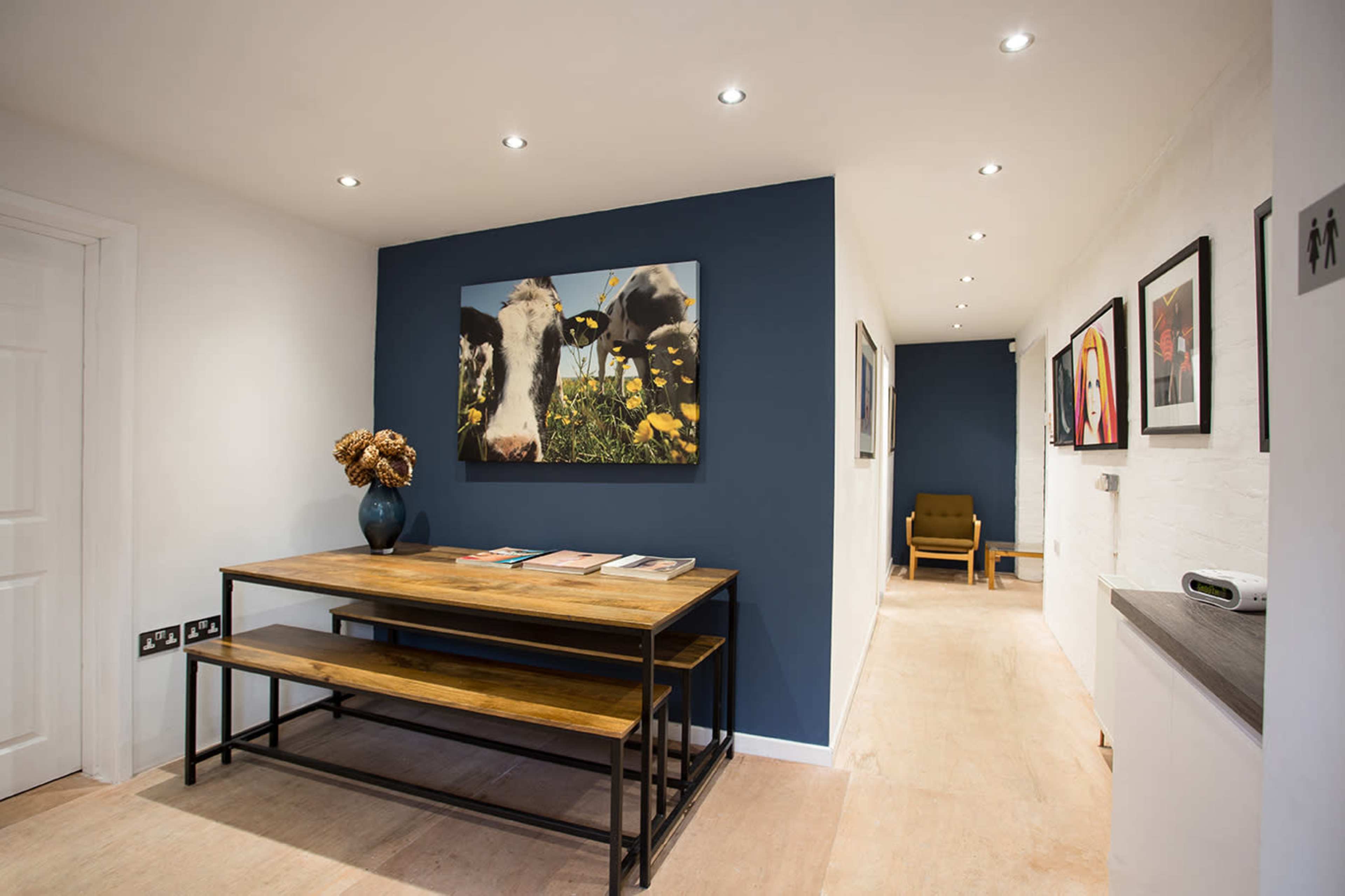 A hallway features a wooden table with magazines and a vase on one side, alongside paintings on the white walls and a blue accent wall with a large cow-themed artwork.