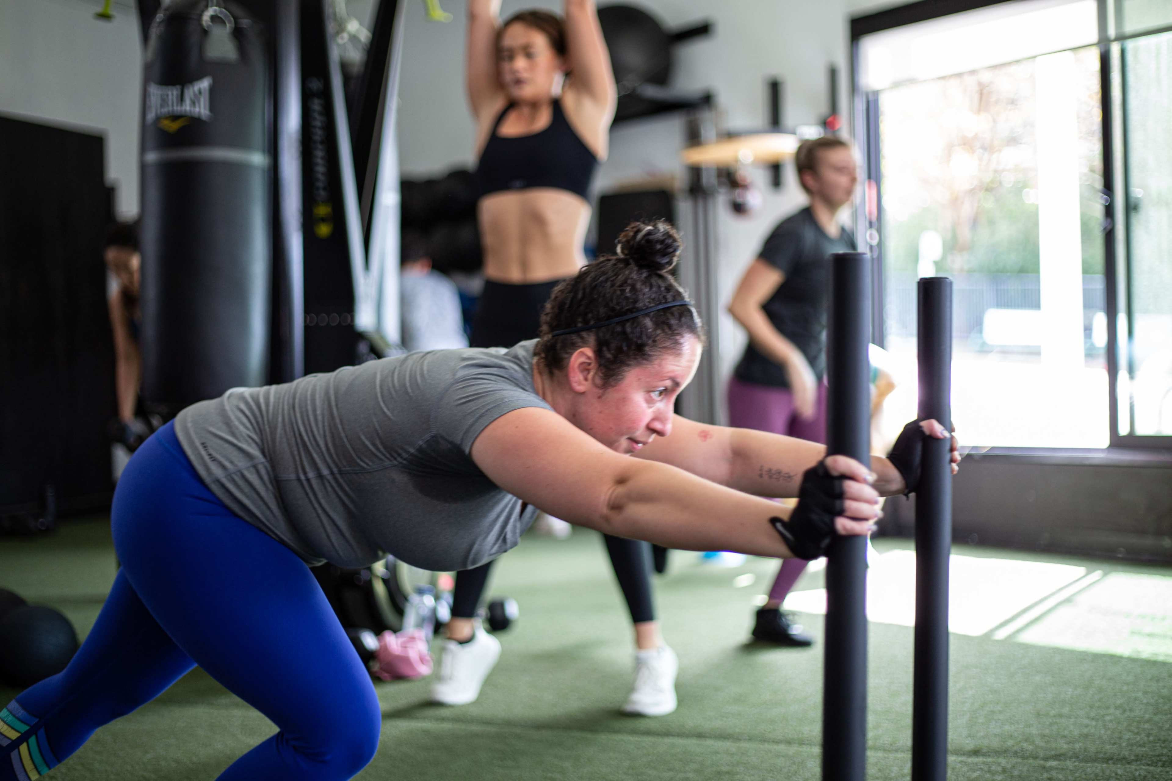A group of people exercises in a gym, with one person pushing a weighted sled while others perform various workouts in the background.