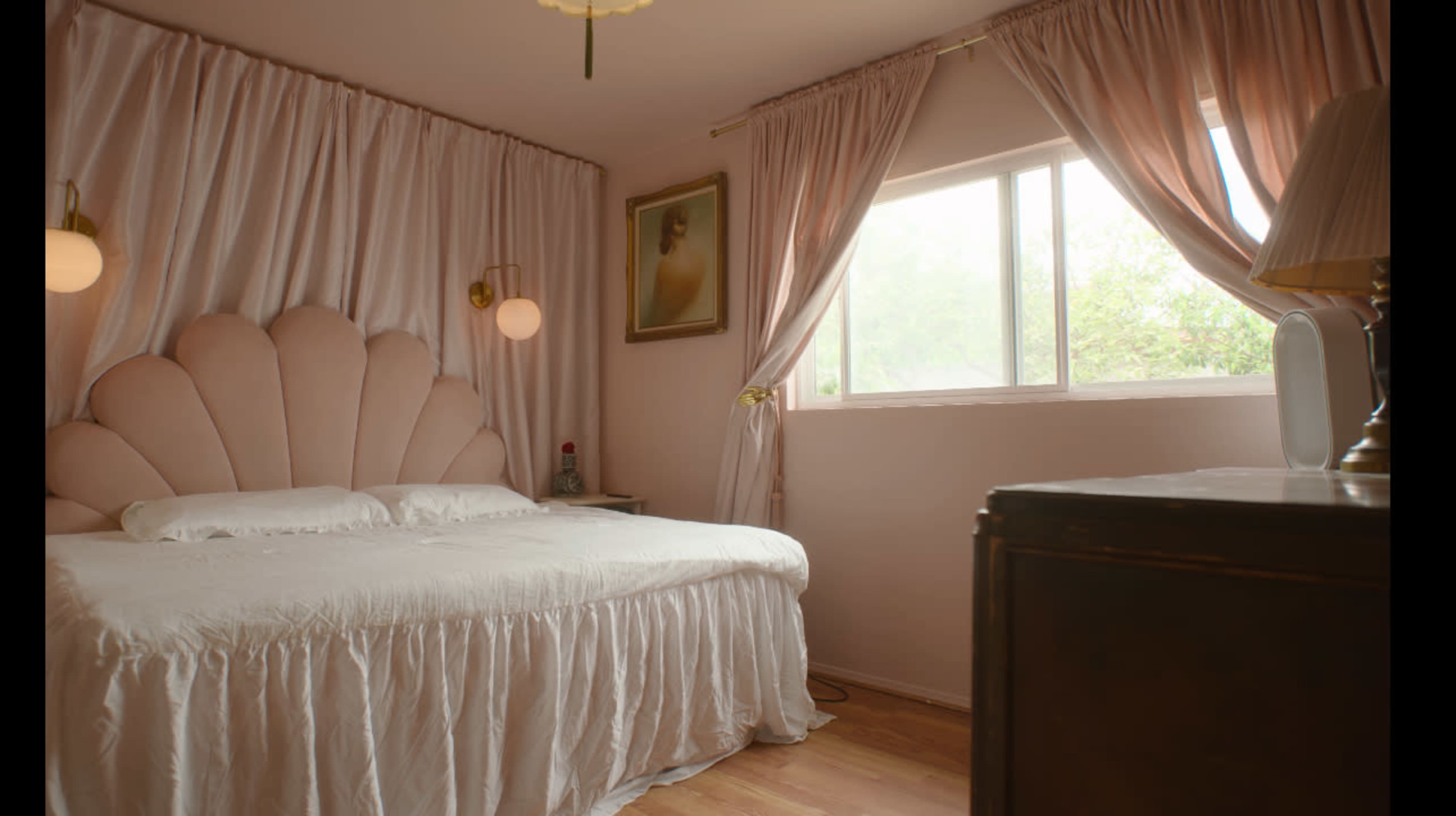 The image shows a softly lit bedroom featuring a large pink upholstered headboard, white bedding, and curtains, with a window allowing natural light to enter.