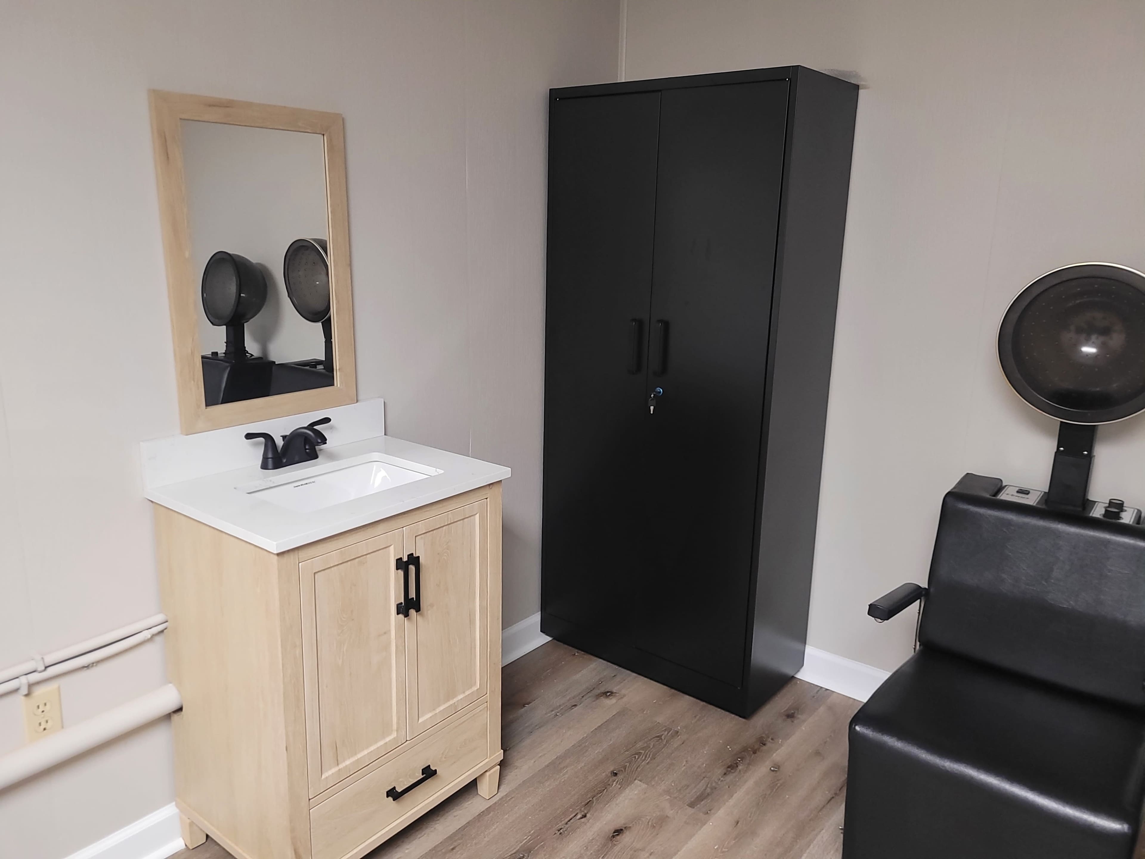 The image shows a small, modern salon space with a wooden vanity and mirror, a black wardrobe, and a styling chair.