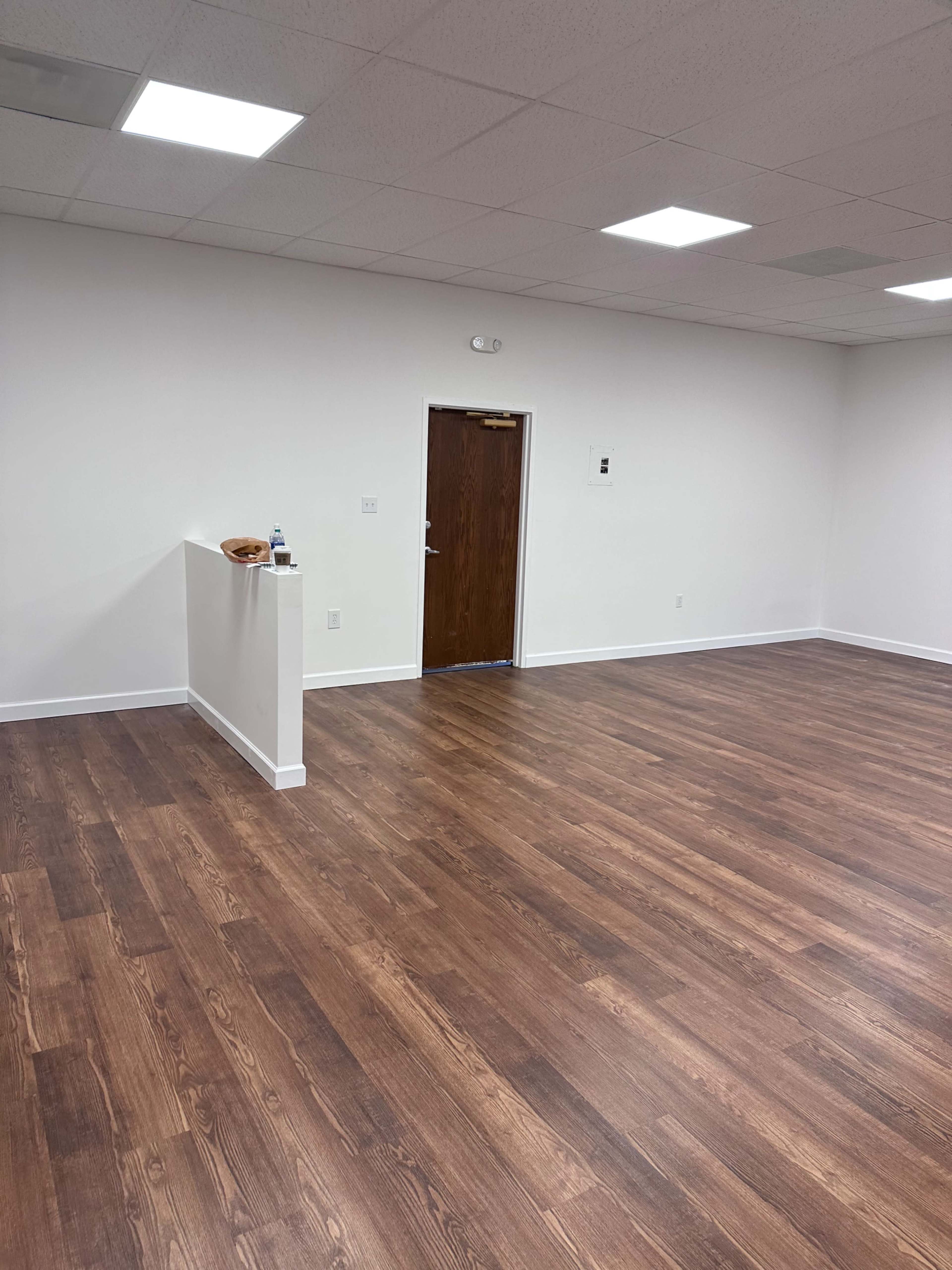 The image shows a spacious, empty room with wooden flooring and white walls, featuring a doorway on one side.