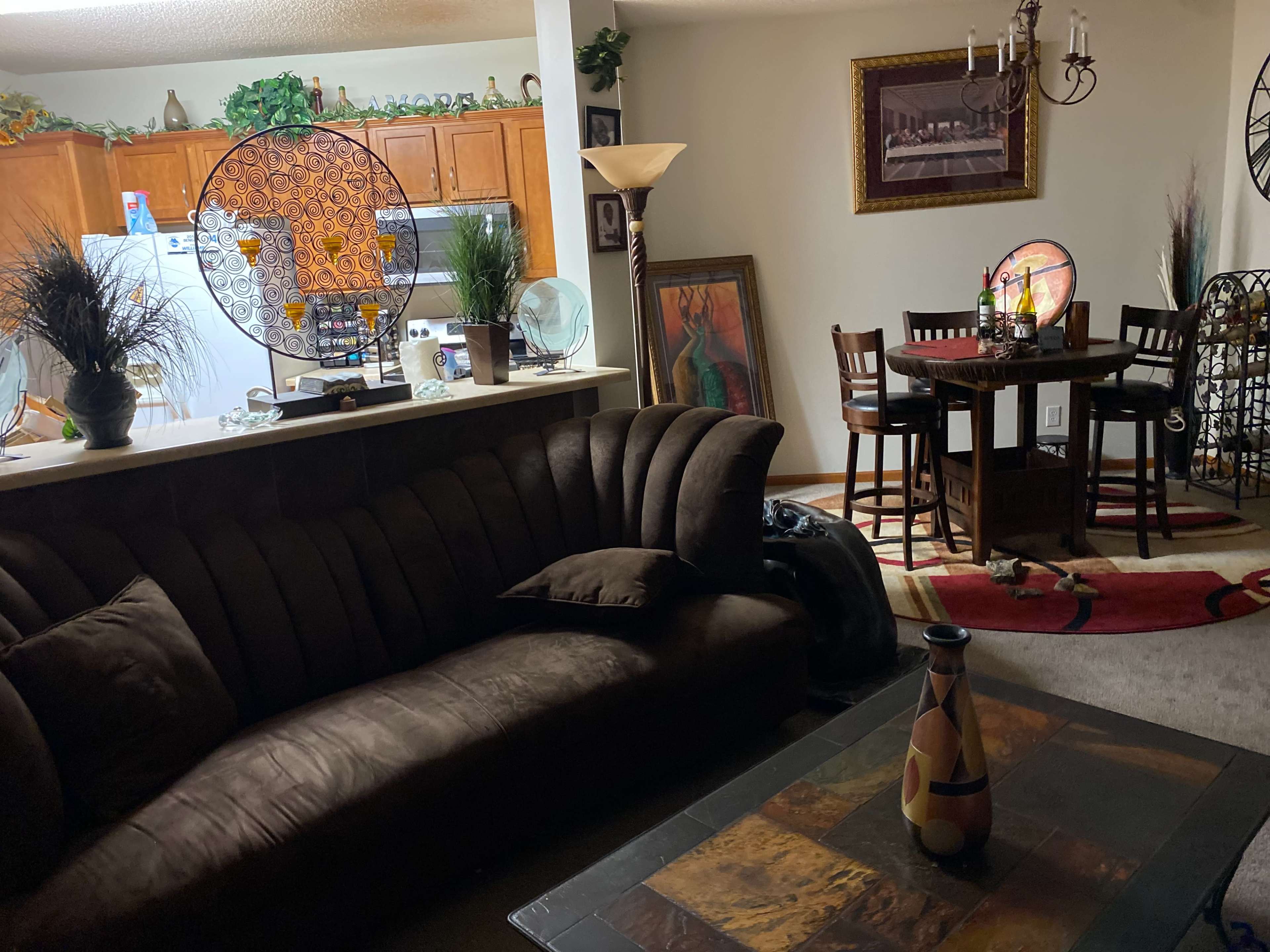 A living room with a brown sofa, a coffee table, a dining area with a small table and chairs, and a kitchen visible in the background.