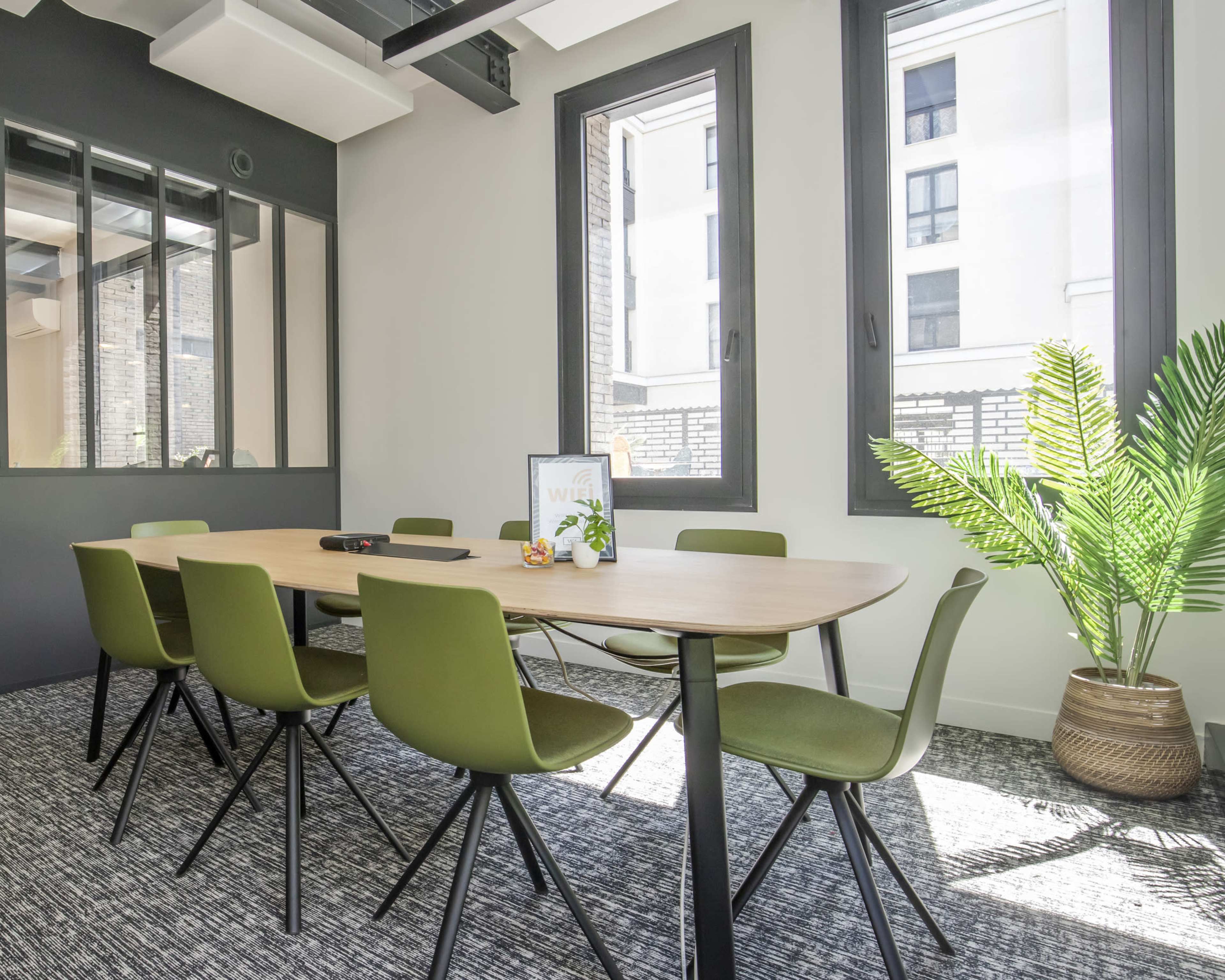 A modern conference room features a long wooden table surrounded by green chairs, with large windows allowing natural light to illuminate the space.