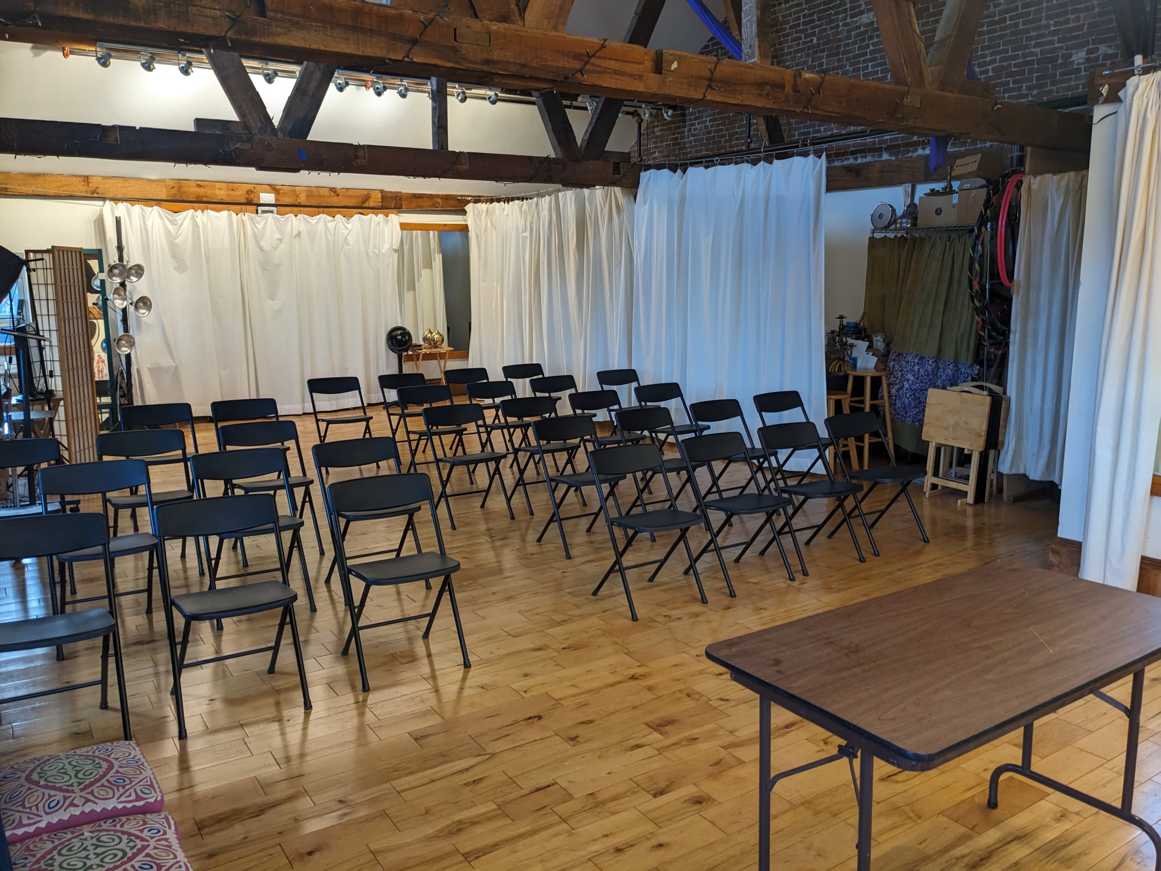 A large room with wooden beams, featuring rows of black folding chairs set up for an event and a table positioned to the side.