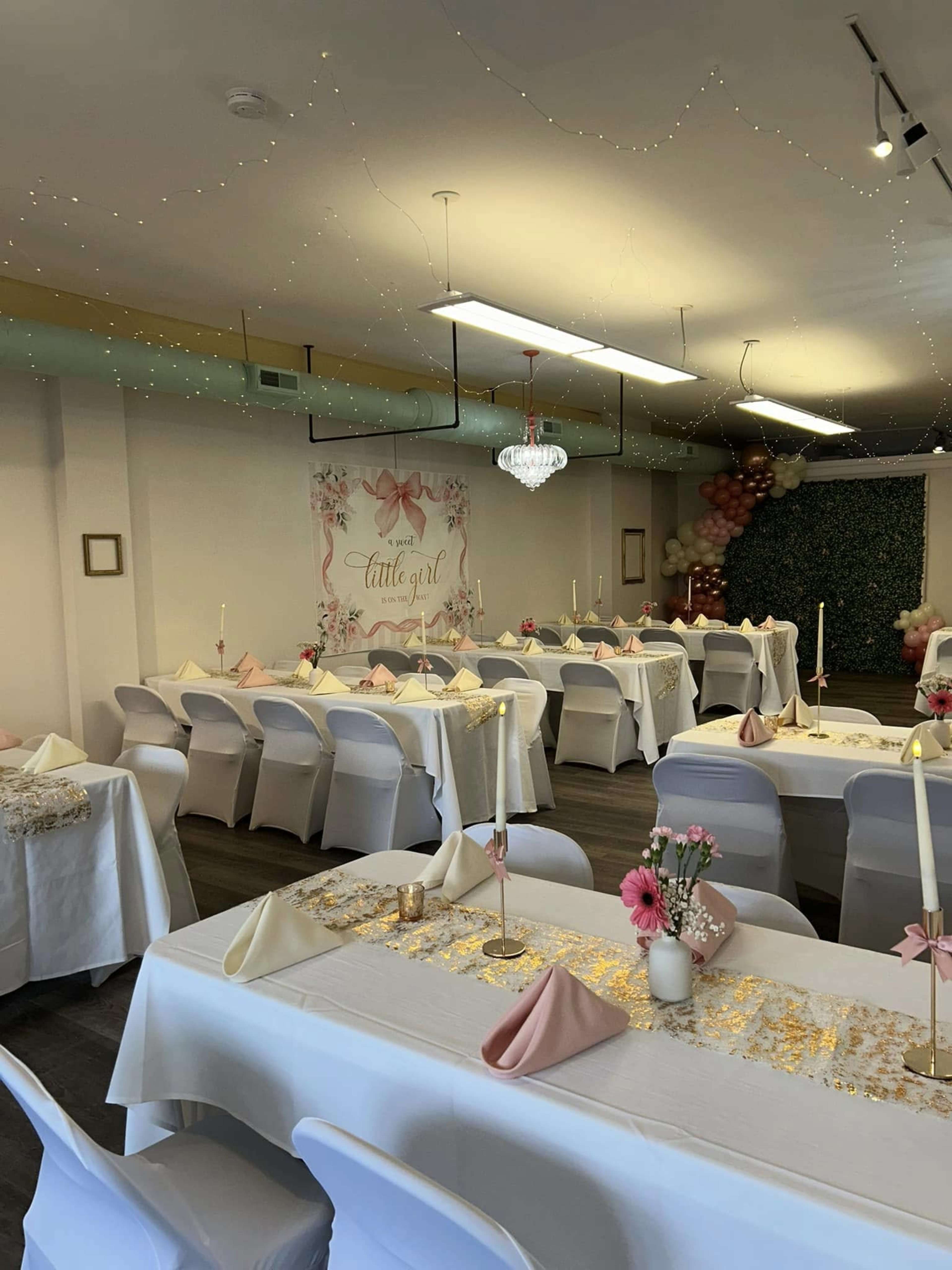 The image shows a decorated event space with neatly arranged tables, each set with white tablecloths and pink napkins, and adorned with candles, flowers, and a backdrop featuring a "Cattle Girl" theme.