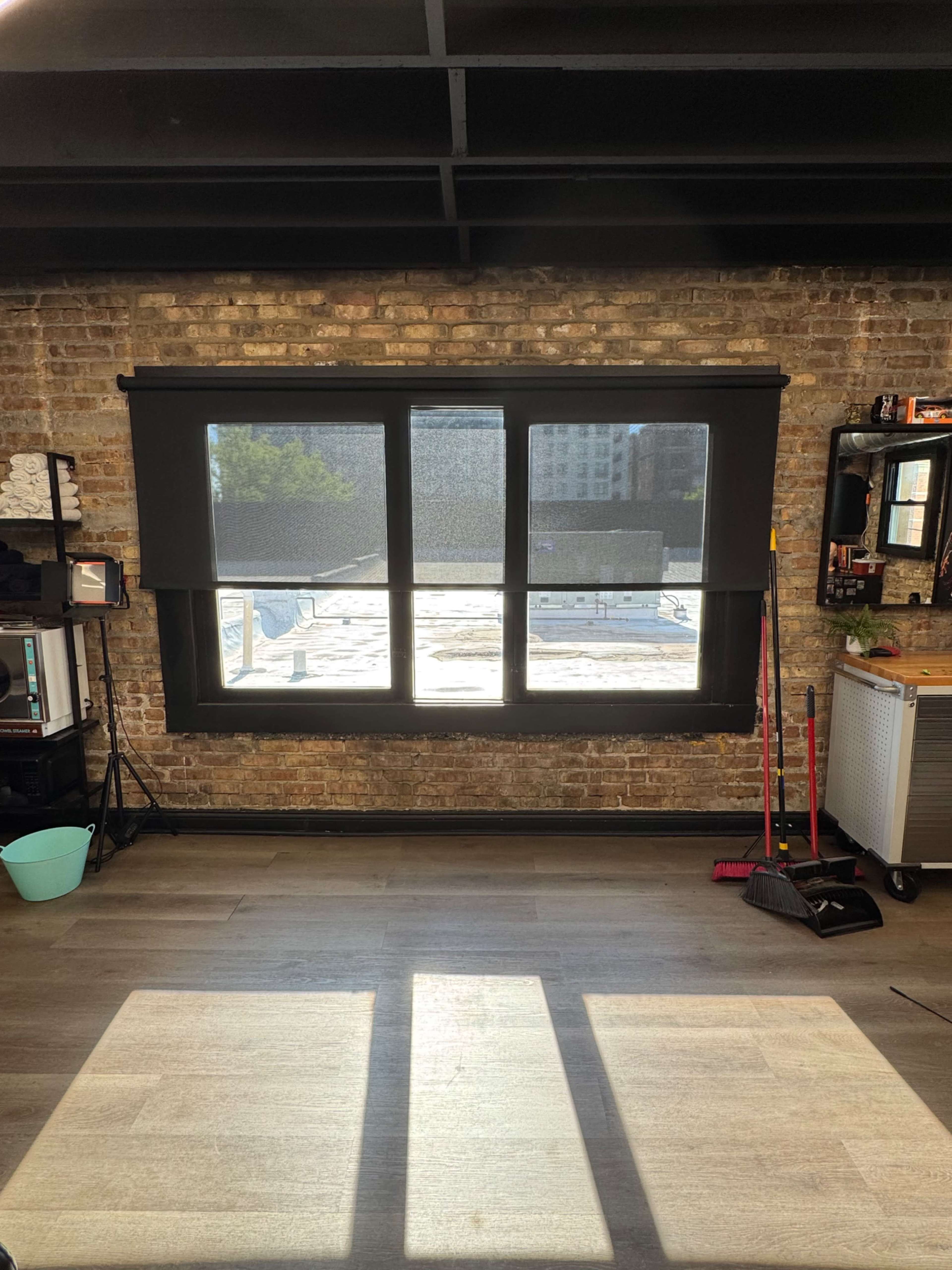 The image shows a large window with black blinds, overlooking a brick wall interior and a wooden floor.