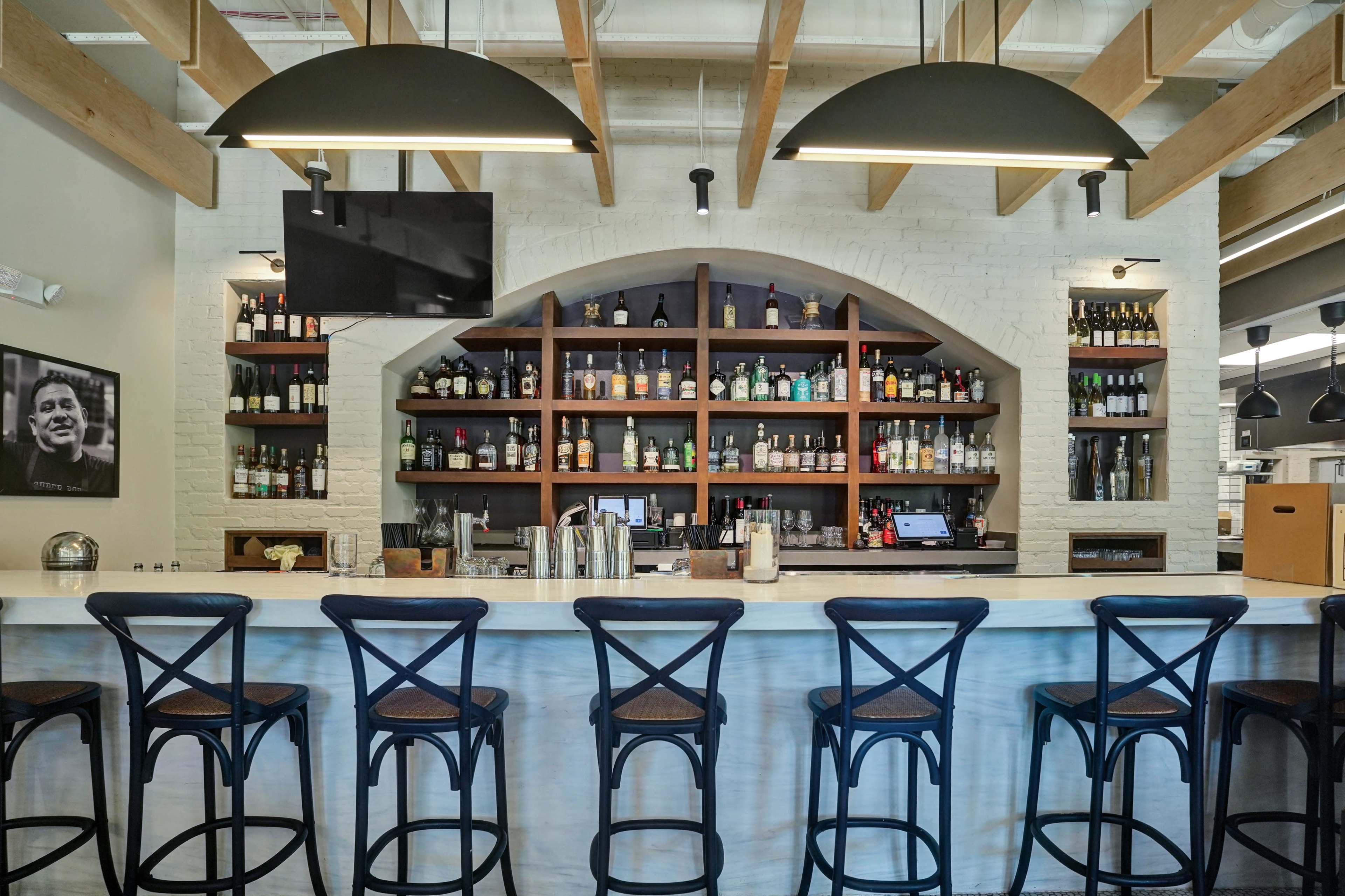 The image features a well-organized bar area with a row of high stools facing a polished counter, surrounded by shelves displaying a variety of liquor bottles and a mounted television.