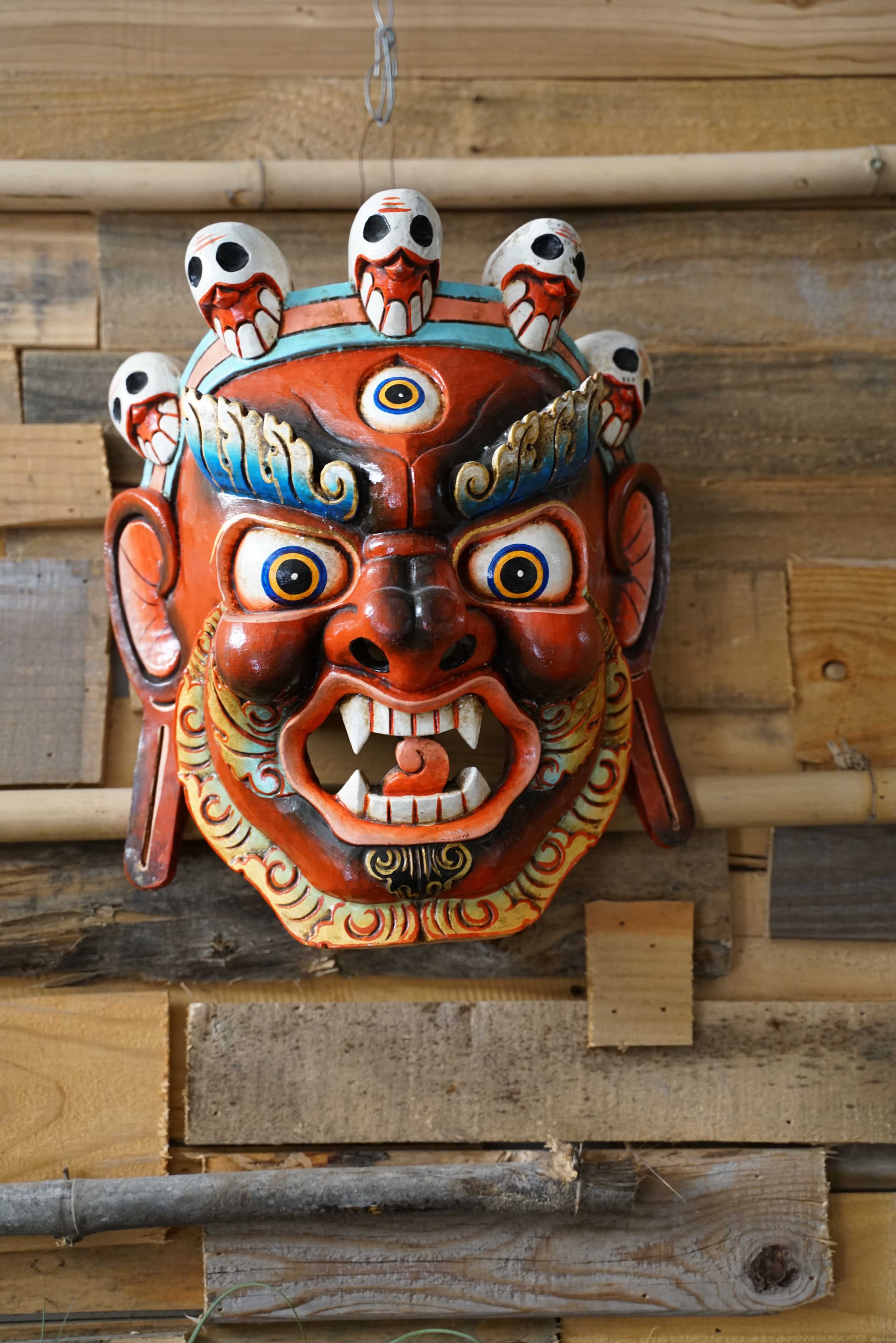 A colorful decorative mask with fierce facial features and multiple eyes is mounted on a wooden wall.
