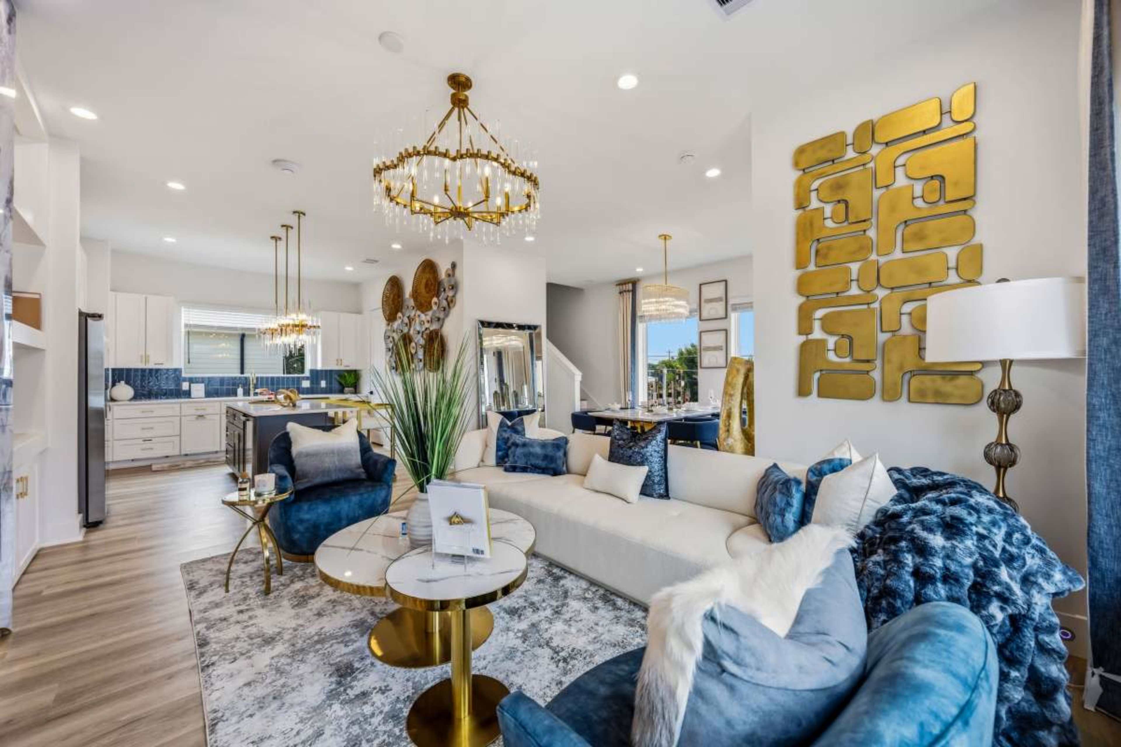 The image shows a modern living room featuring a large white sofa, blue accent chairs, a round coffee table, a decorative wall with gold elements, and chandeliers above.