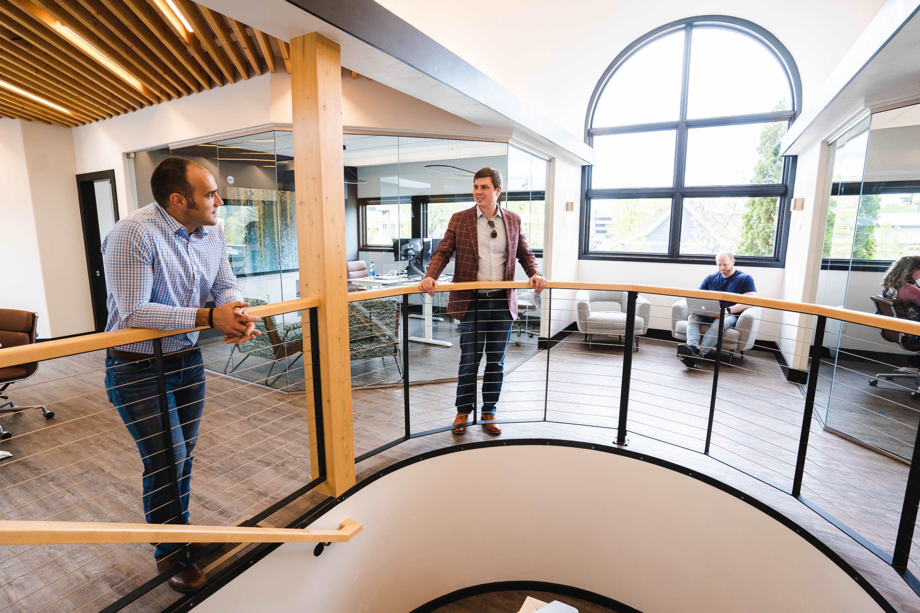 Two men converse while leaning on a wooden railing above a circular staircase in a modern office space, with a glass-walled meeting area in the background.