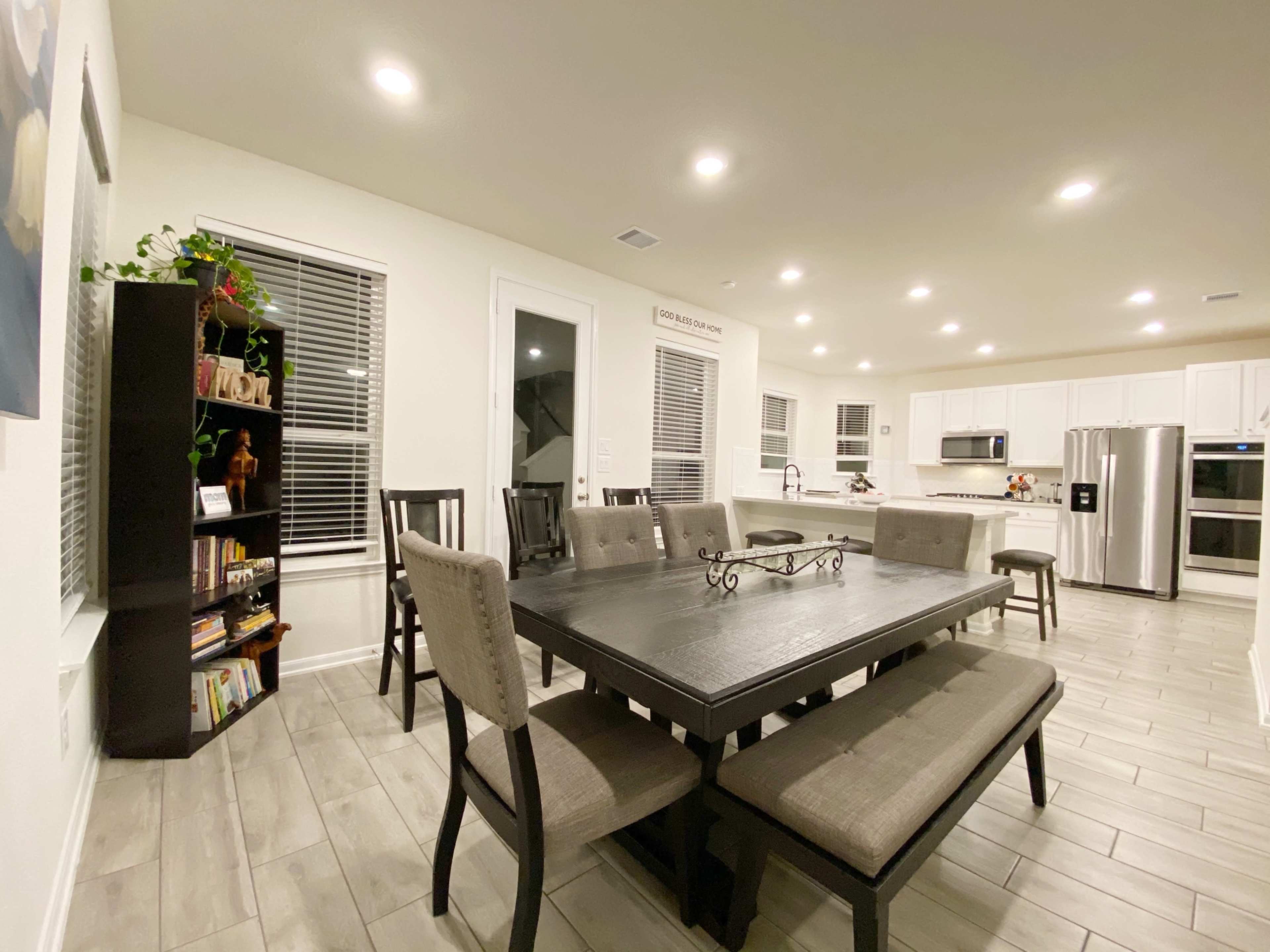 A spacious dining area features a rectangular black table surrounded by beige upholstered chairs, with a modern kitchen visible in the background.
