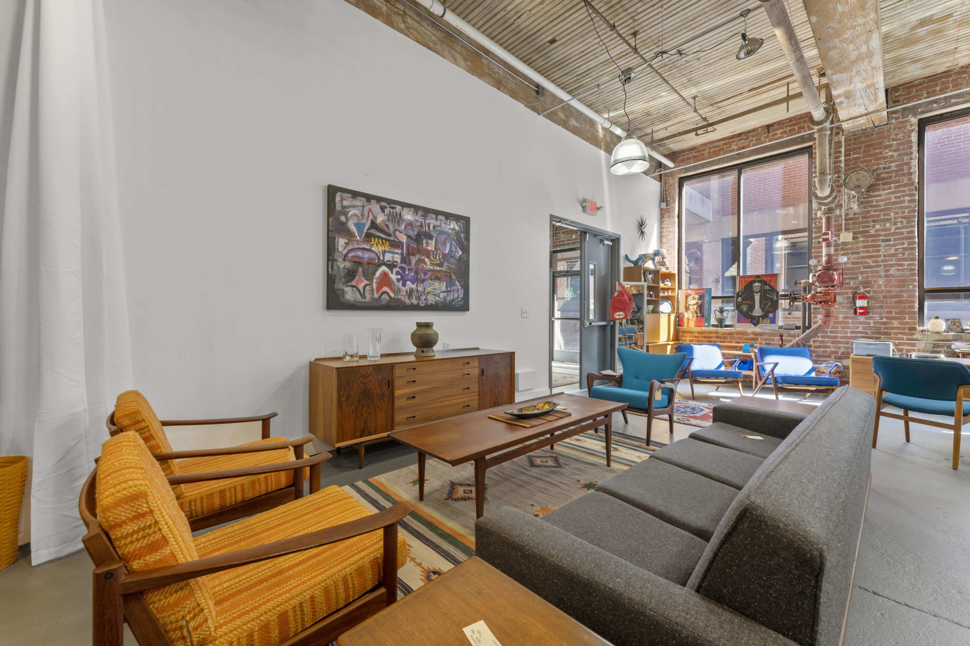 The image shows a modern living space with mid-century furniture, including a gray sofa and wooden chairs, set against exposed brick walls and large windows.