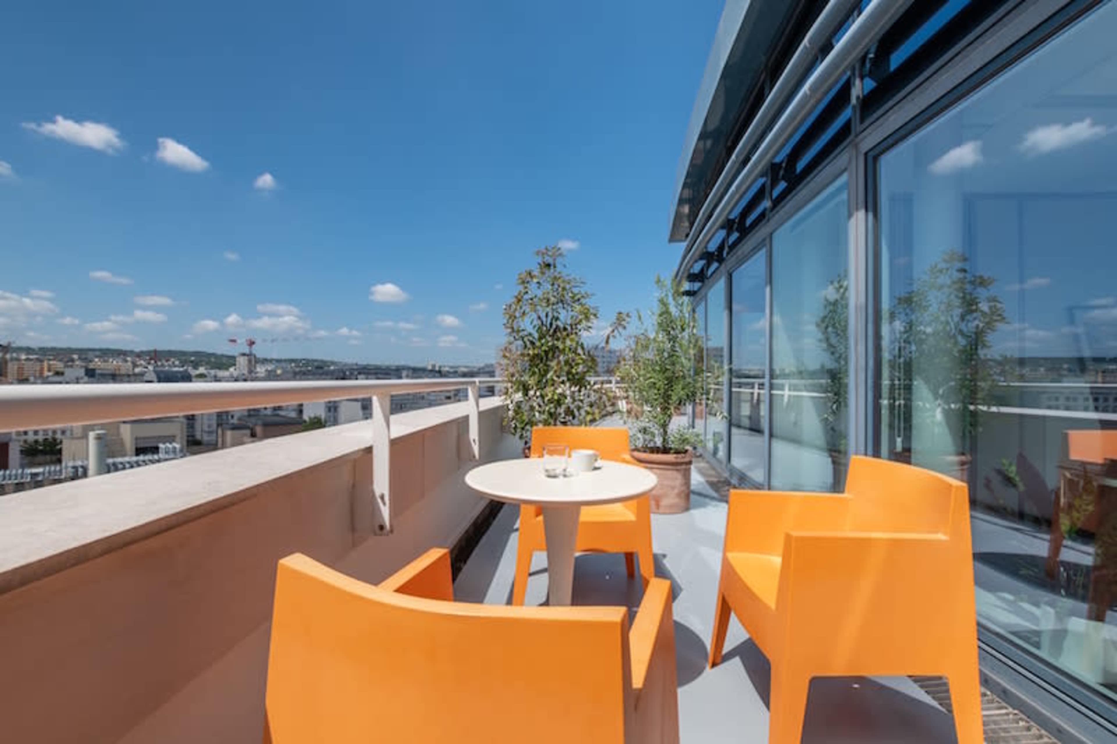 The image shows a rooftop terrace with orange chairs and a small table, surrounded by potted plants and a clear blue sky.