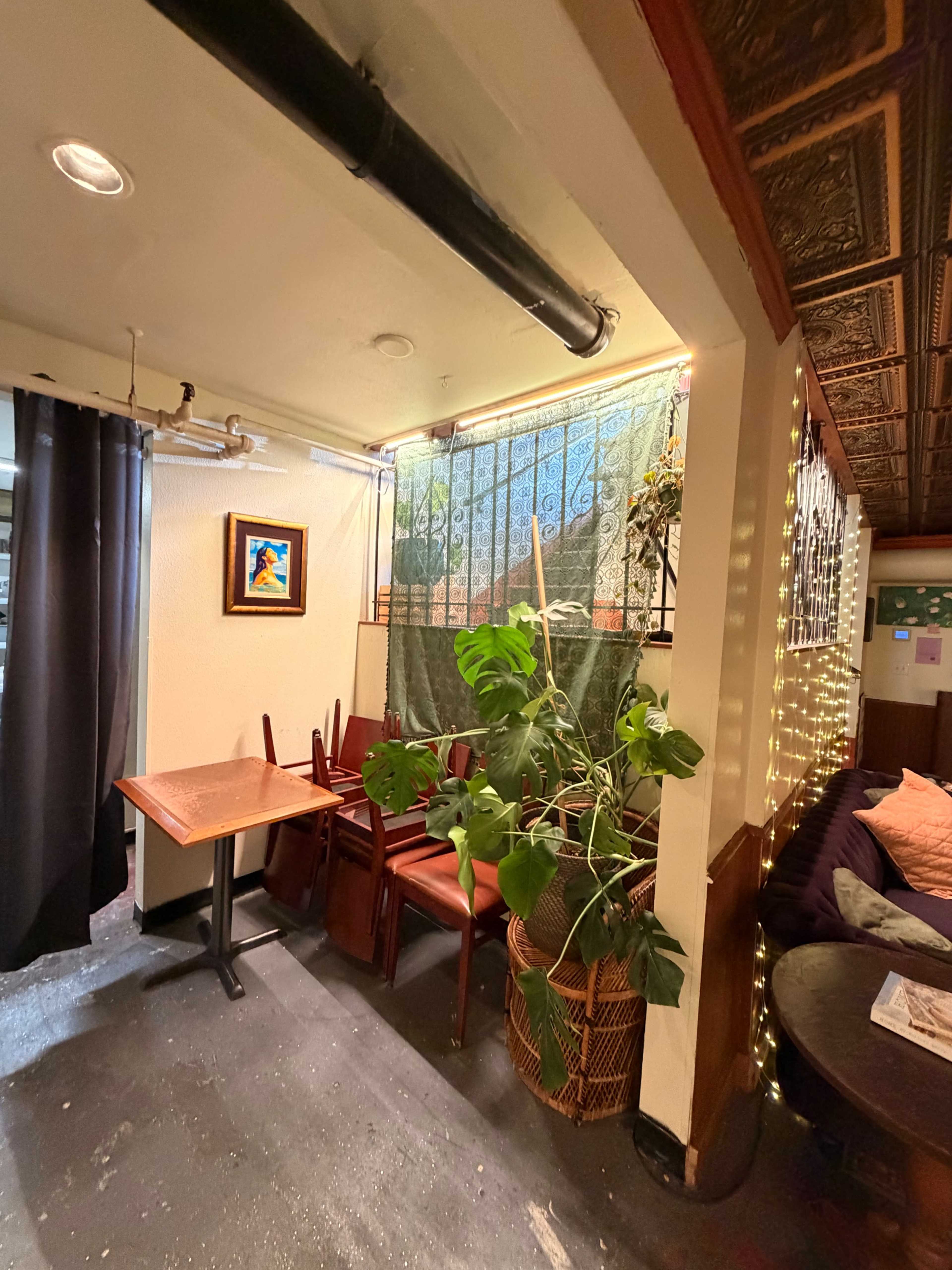 The image shows a cozy corner of a cafe with stacked chairs on a table, plants, a painted artwork on the wall, and warm lighting decorations.