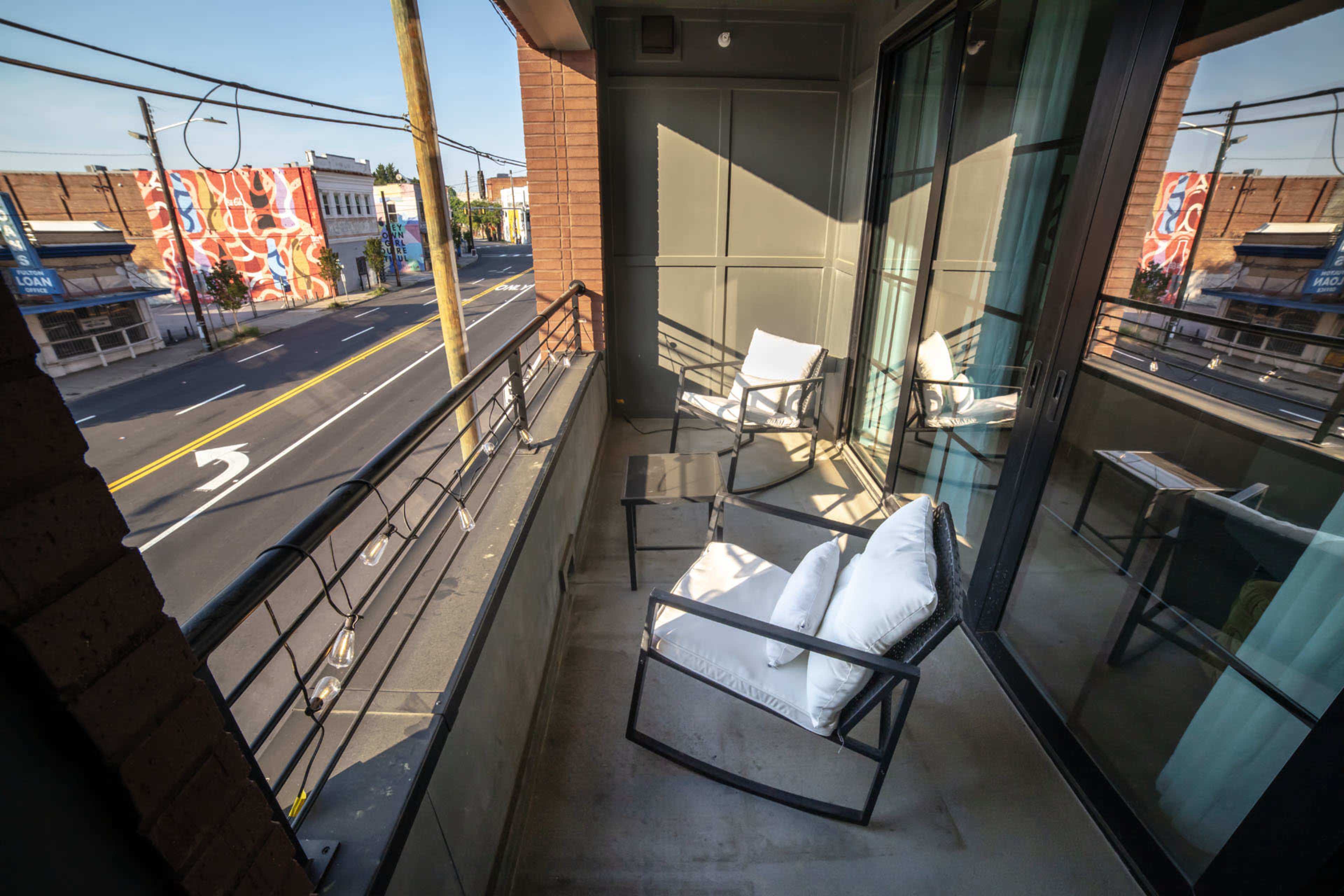 The image shows a balcony with two white cushioned chairs overlooking a street featuring colorful wall art.