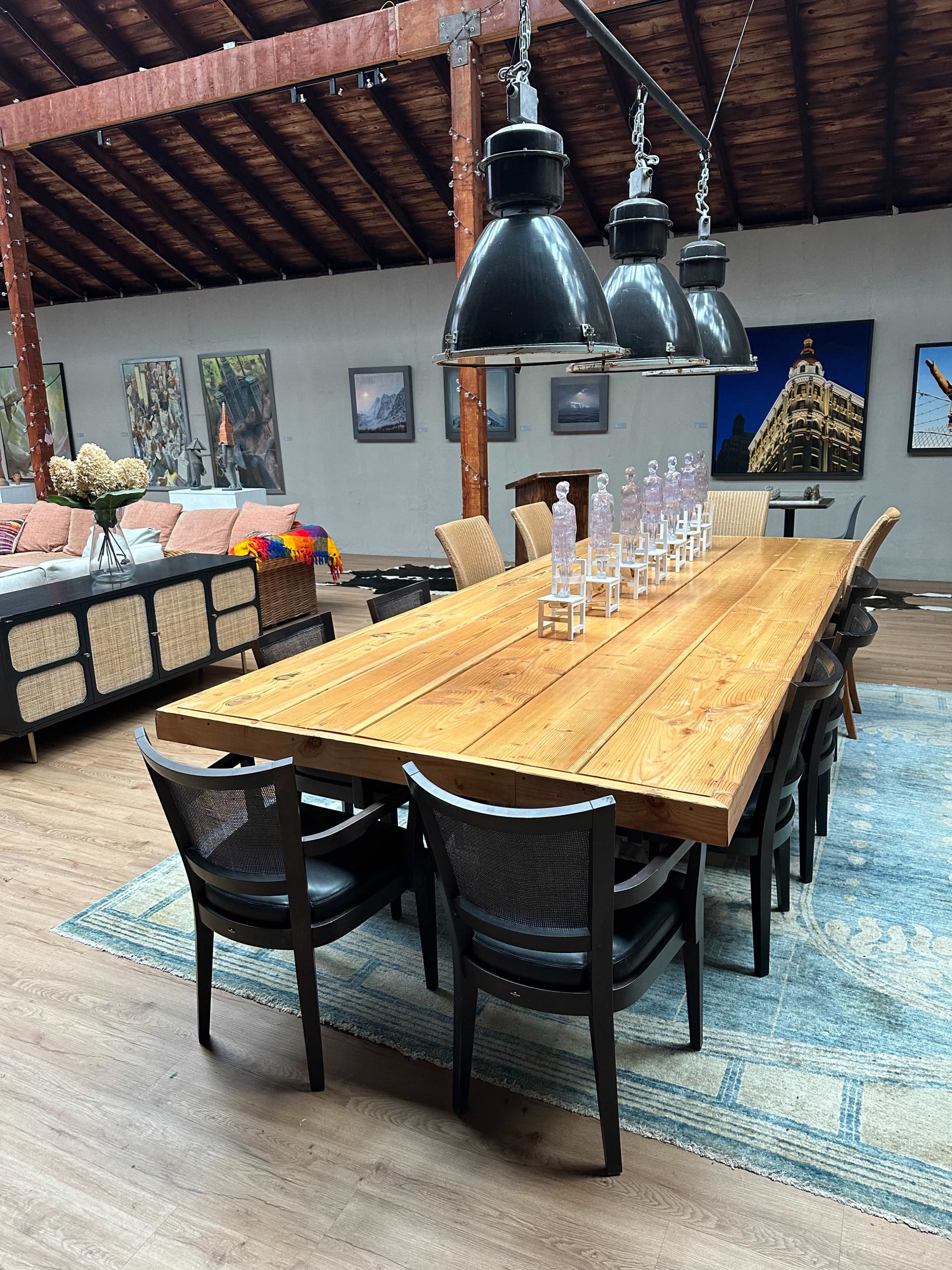 A long wooden dining table is centered in a spacious room, surrounded by black chairs, with large pendant lights hanging above and various artworks on the walls.