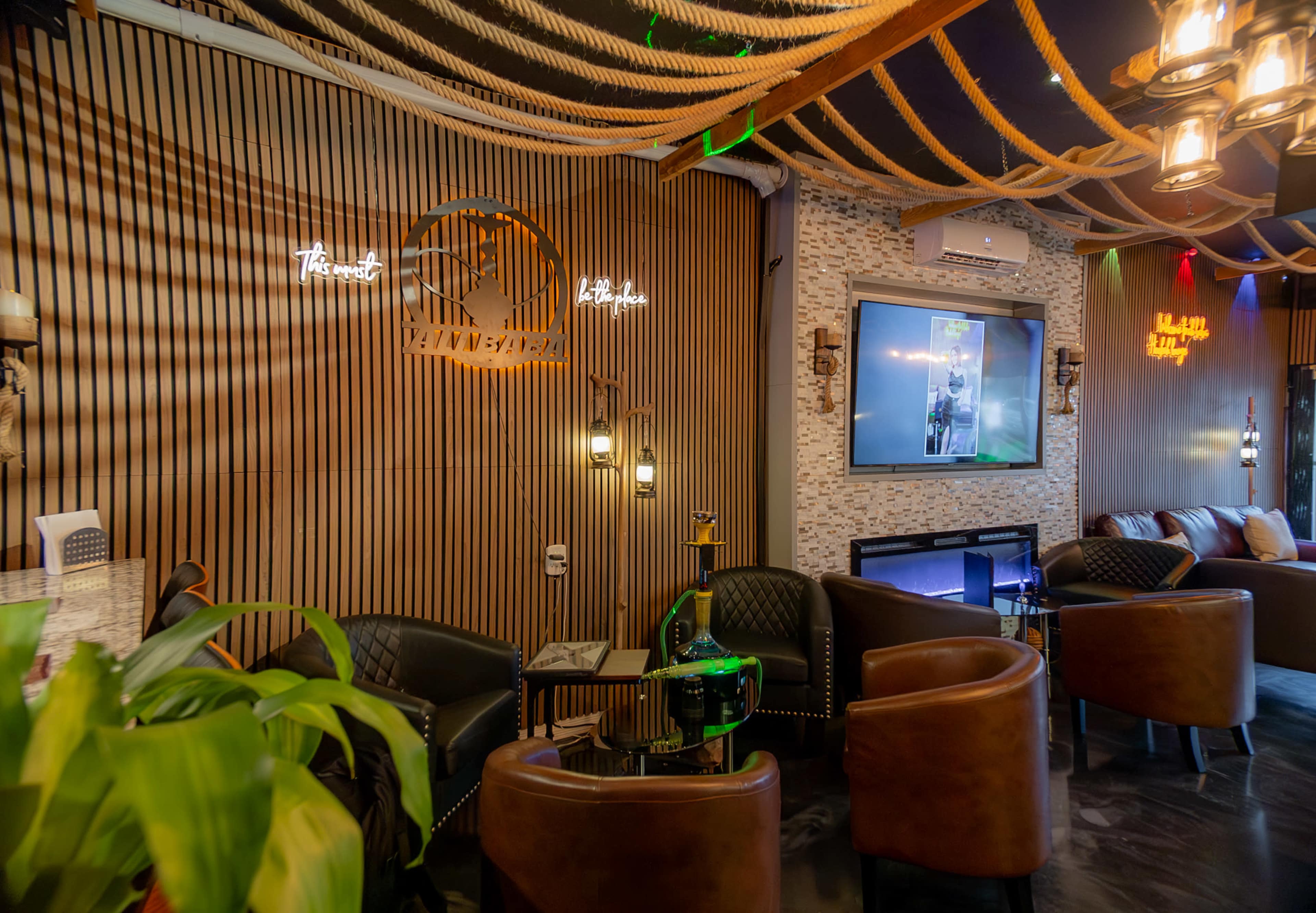 A modern lounge area with wooden paneling, black leather chairs, decorative lighting, and a television mounted on the wall.