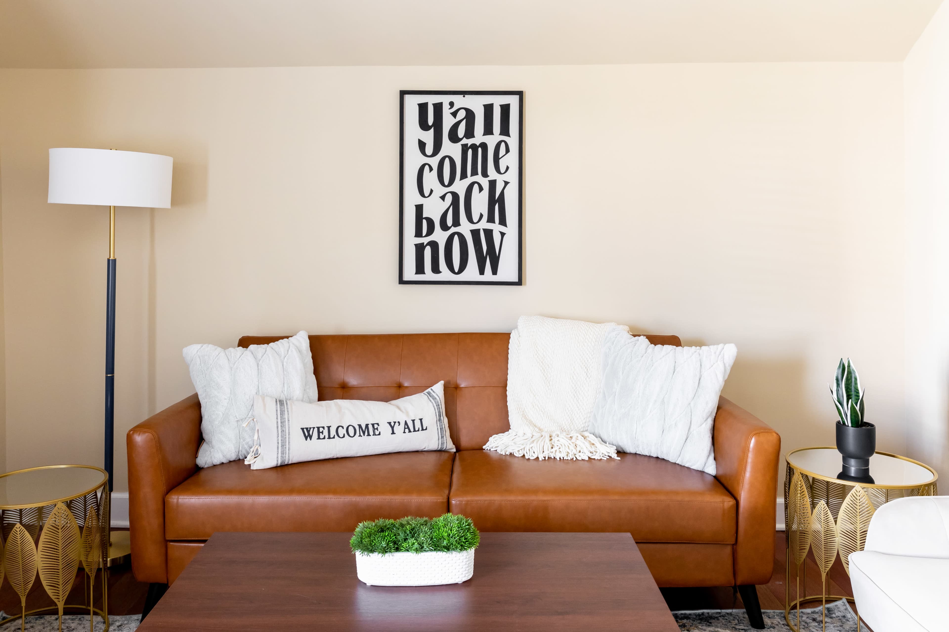 A brown leather sofa with white pillows and a decorative throw sits in a living room, accompanied by side tables and a framed wall art above.