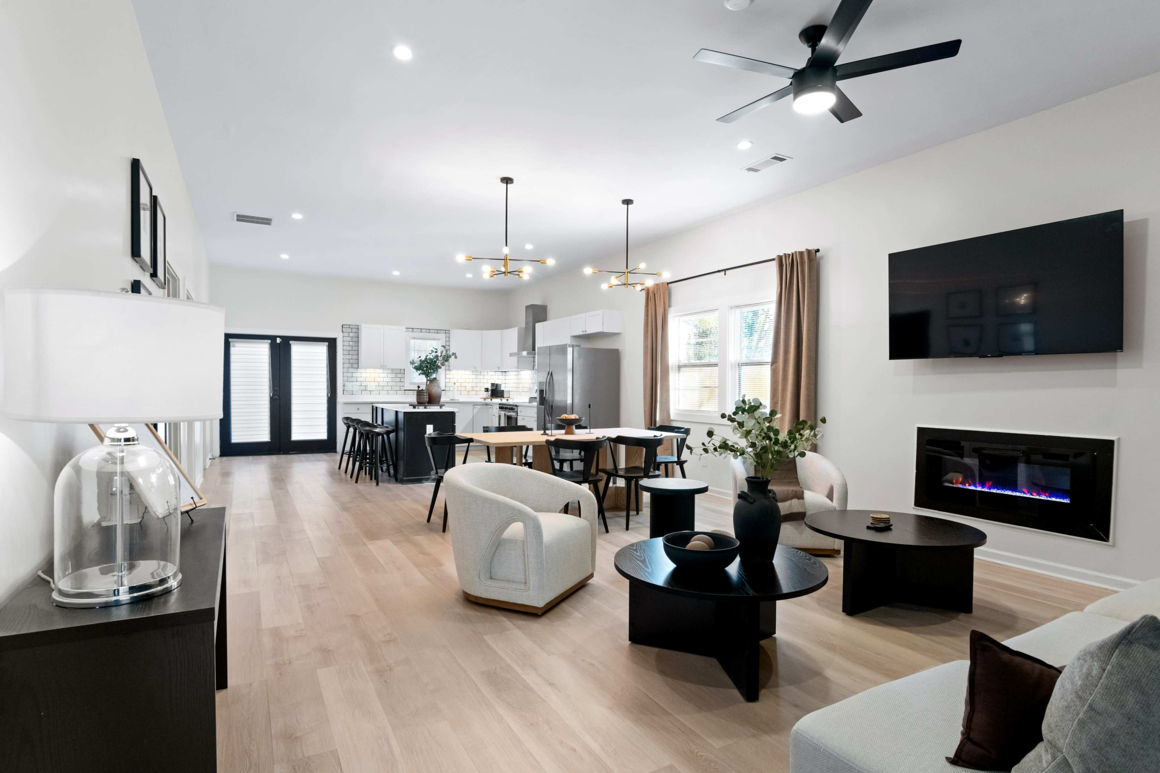 The image depicts a modern open-concept living space featuring a kitchen, dining area, and lounge, characterized by minimalist furniture and large windows.