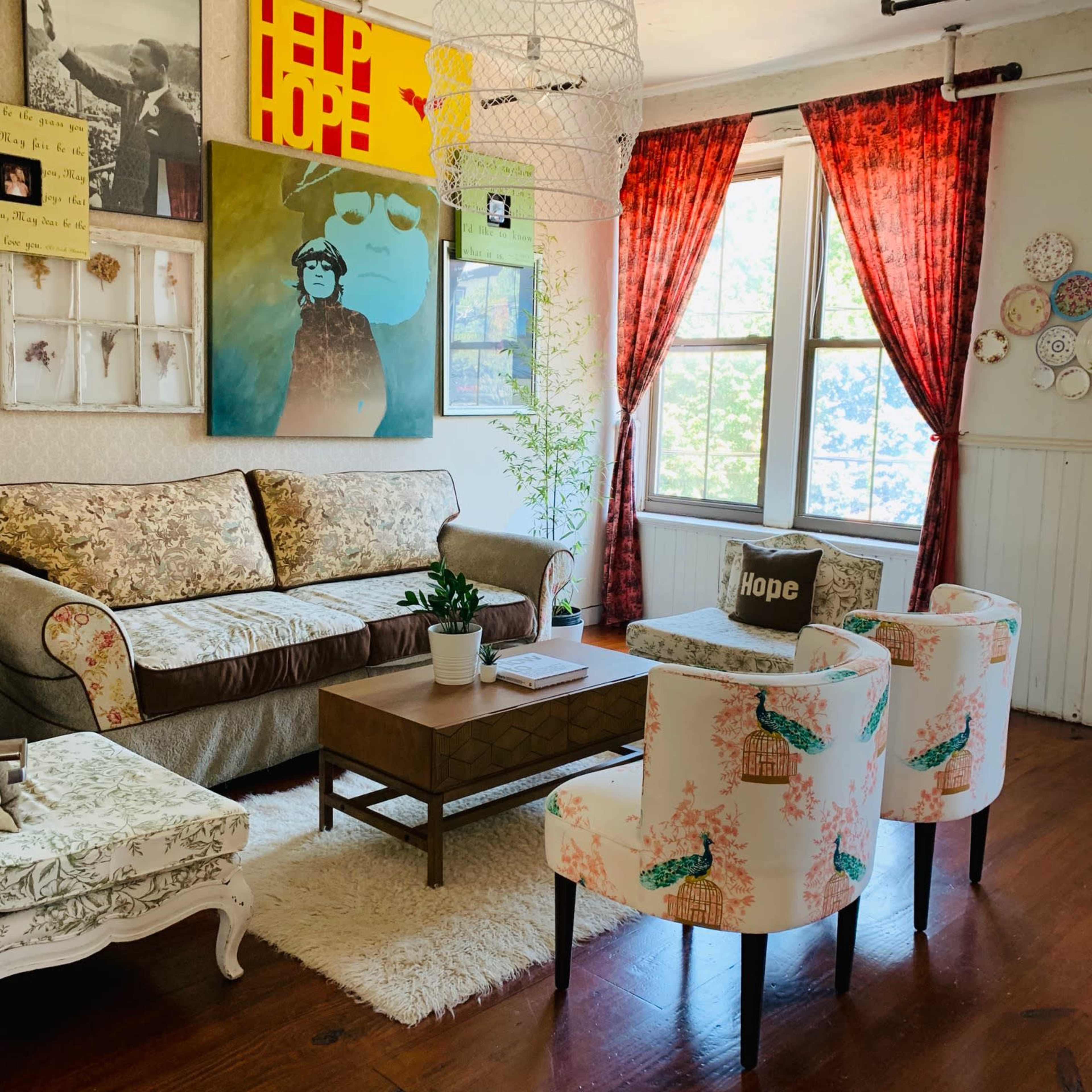A cozy living room features a patterned sofa, a wooden coffee table, and four chairs, with colorful artwork and decor on the walls and a large window letting in natural light.