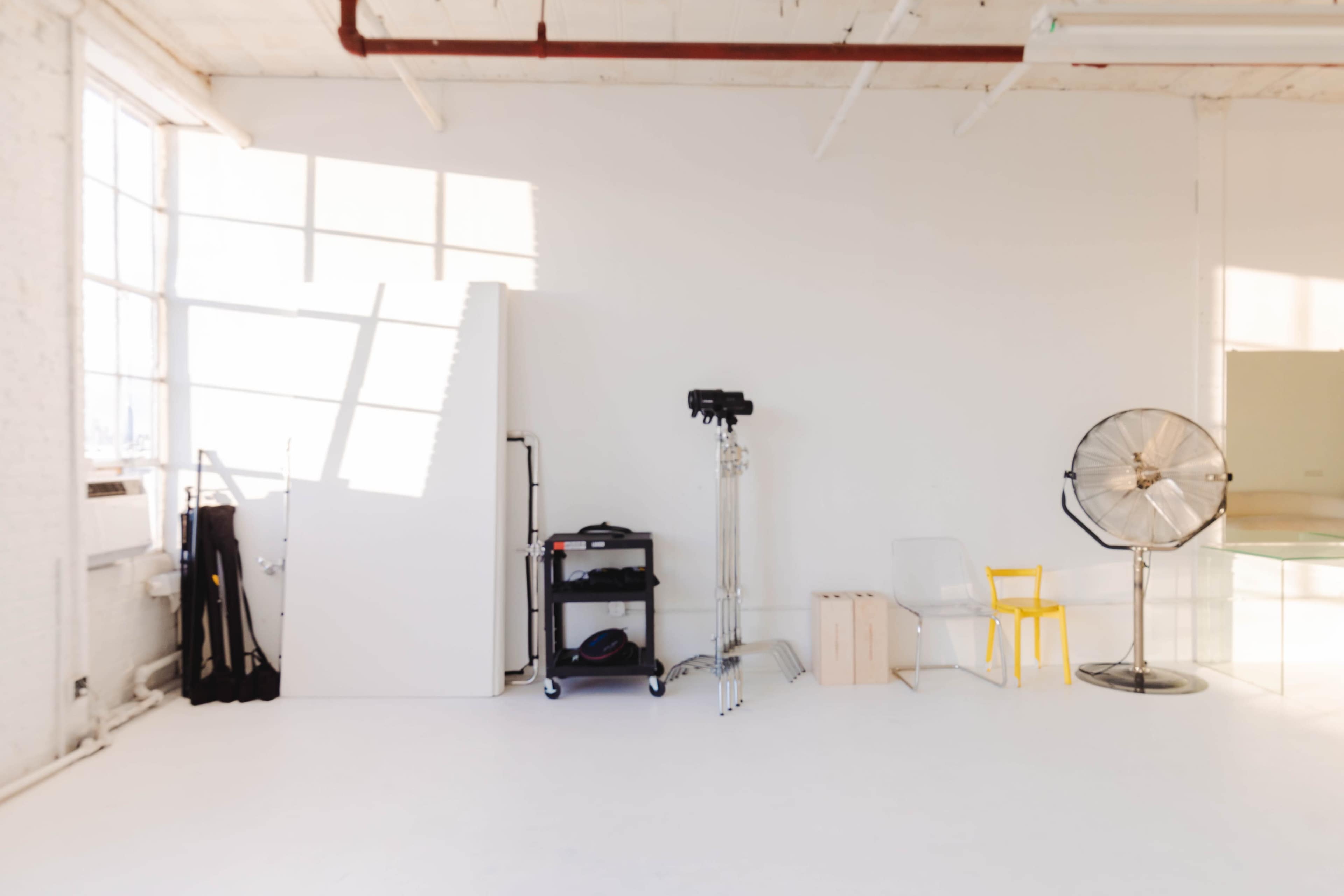 A bright, minimalist studio space displays a white wall, a yellow chair, and various equipment, including a fan and stands.