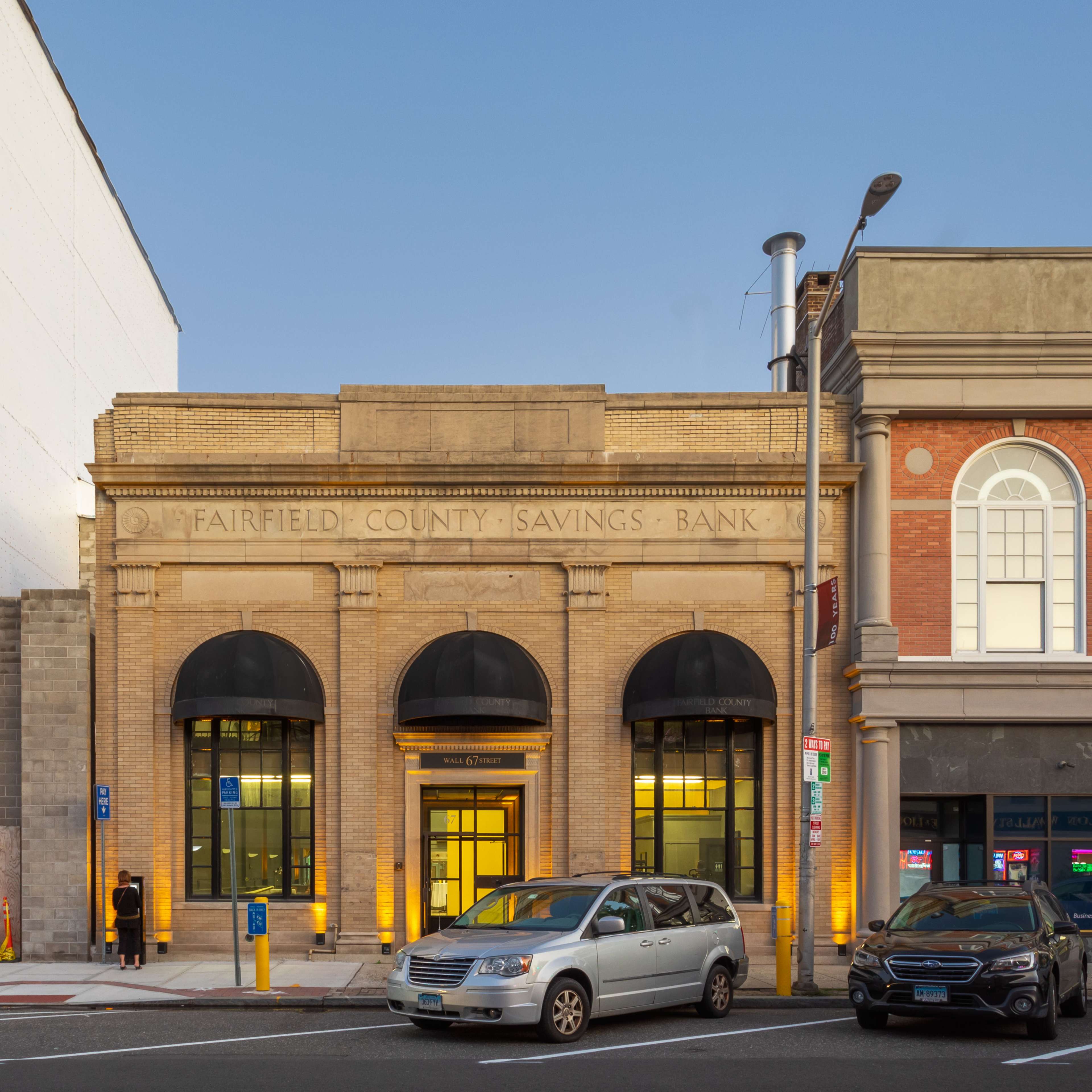 The image shows the facade of the Fairfield County Savings Bank, featuring a symmetrical design with large windows and awnings, positioned next to a brick building.