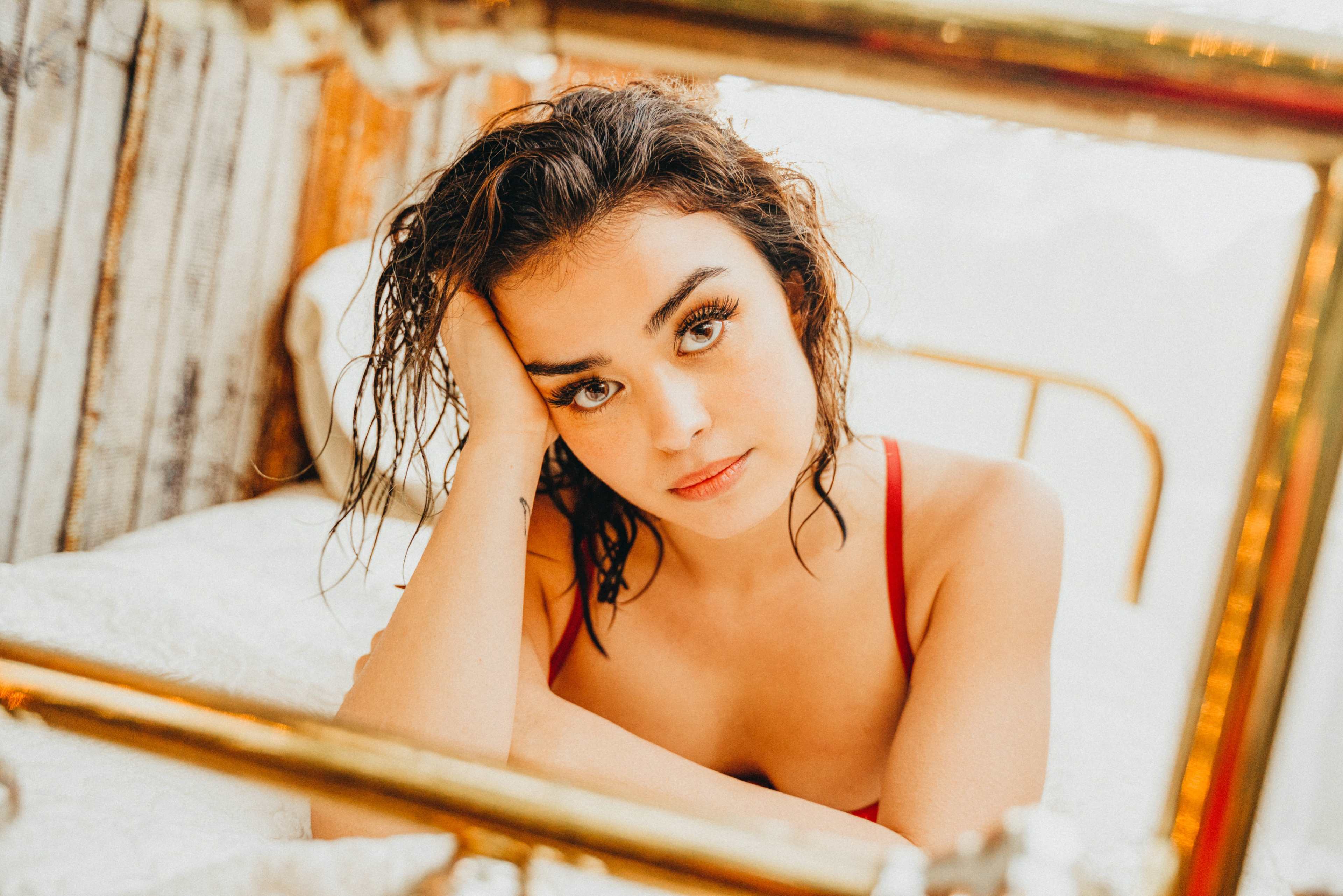 A young woman with wet hair rests her head on her hand while looking directly at the camera from a bed with a vintage frame.