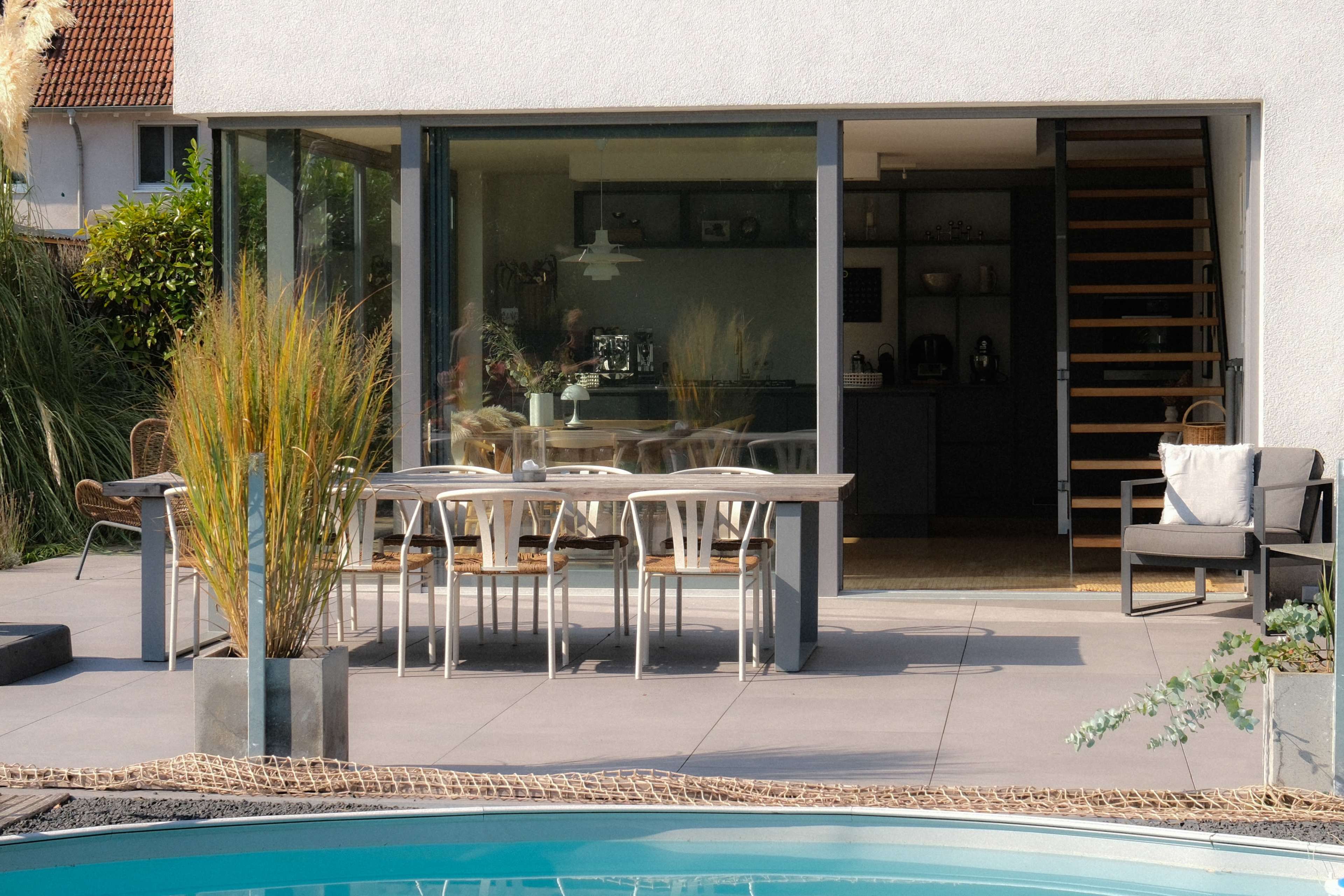 The scene features a modern outdoor dining area with a table and chairs adjacent to a pool, surrounded by greenery and a contemporary home.