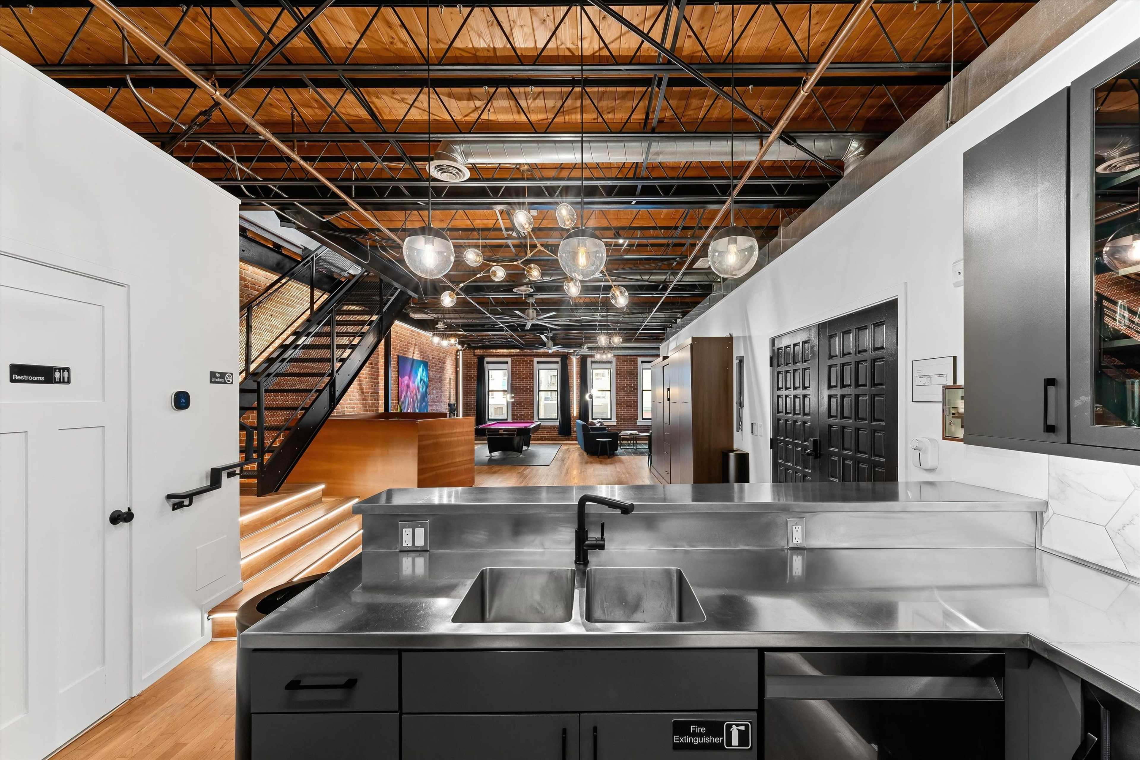 The image shows a modern kitchen area with stainless steel fixtures, leading into a spacious open-concept living space featuring wooden floors and industrial-style lighting.